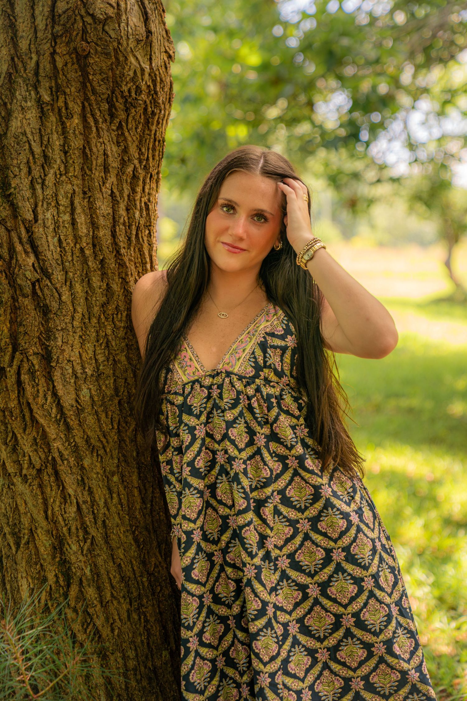 Young woman in floral sundress leaning against tree trunk in lush green park, hand in long dark hair.