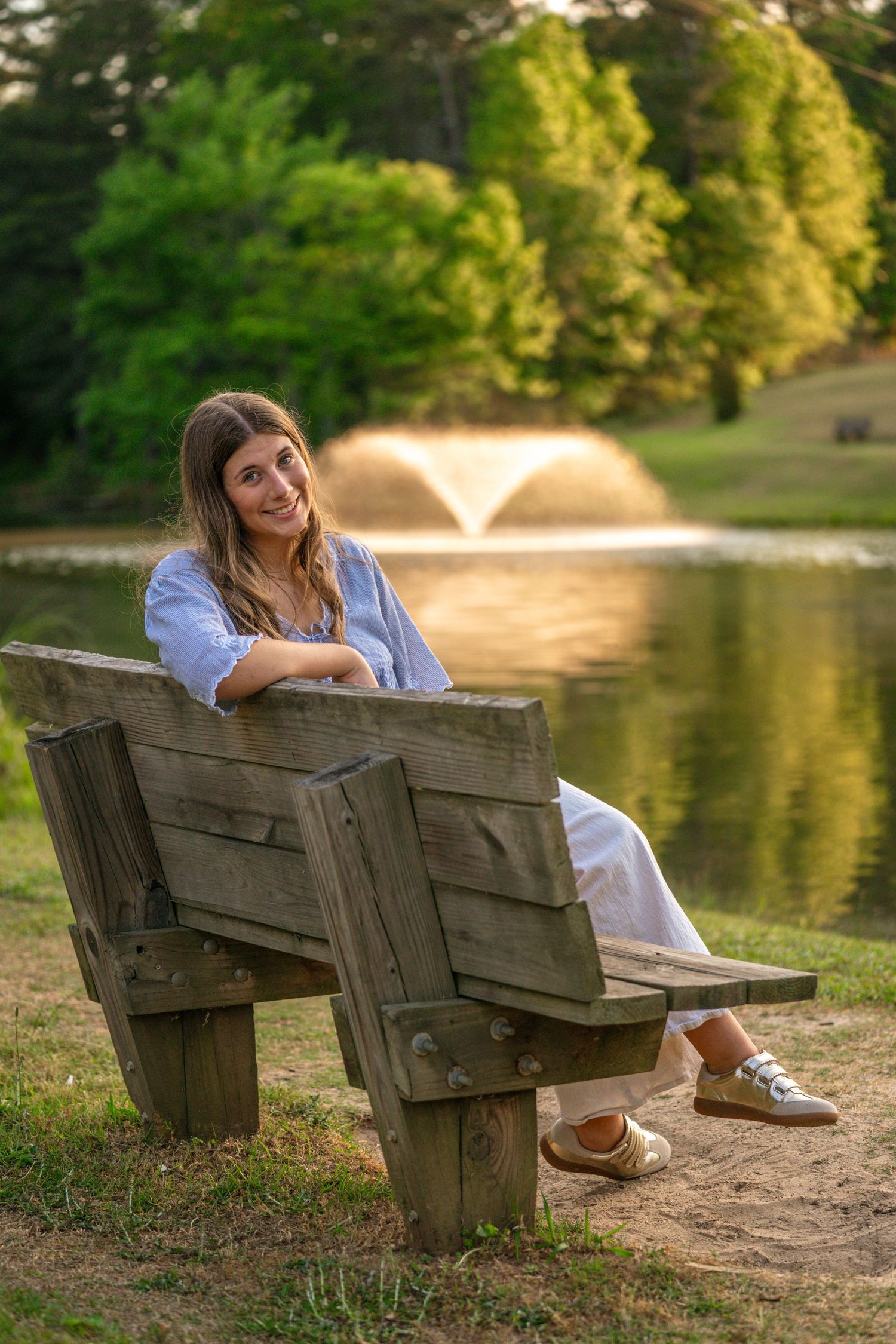 Young woman sitting relaxed on a wooden bench by a serene pond with a fountain and lush green trees at sunset.