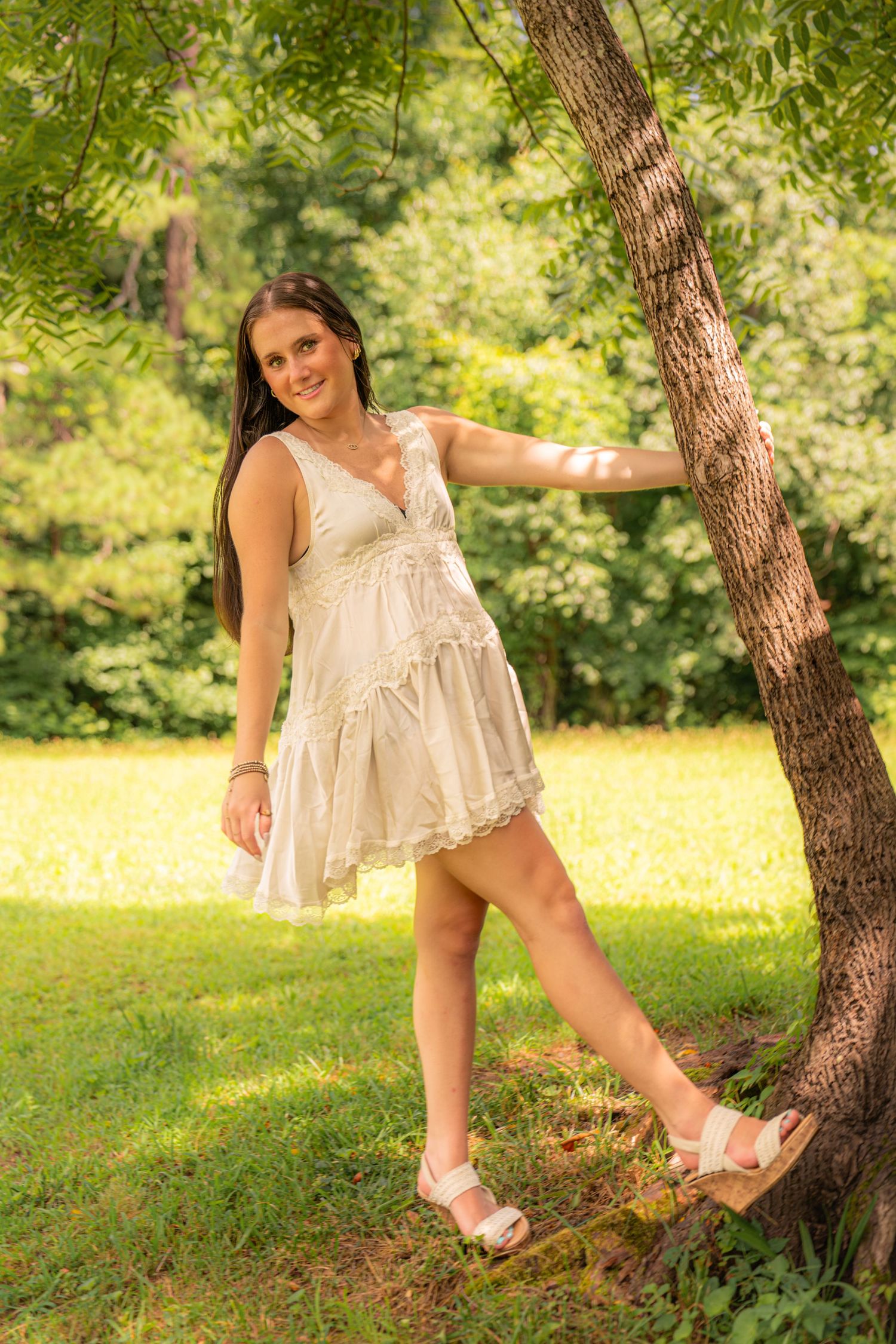 Young woman in white sundress leaning against tree in sunny green park, smiling outdoors.