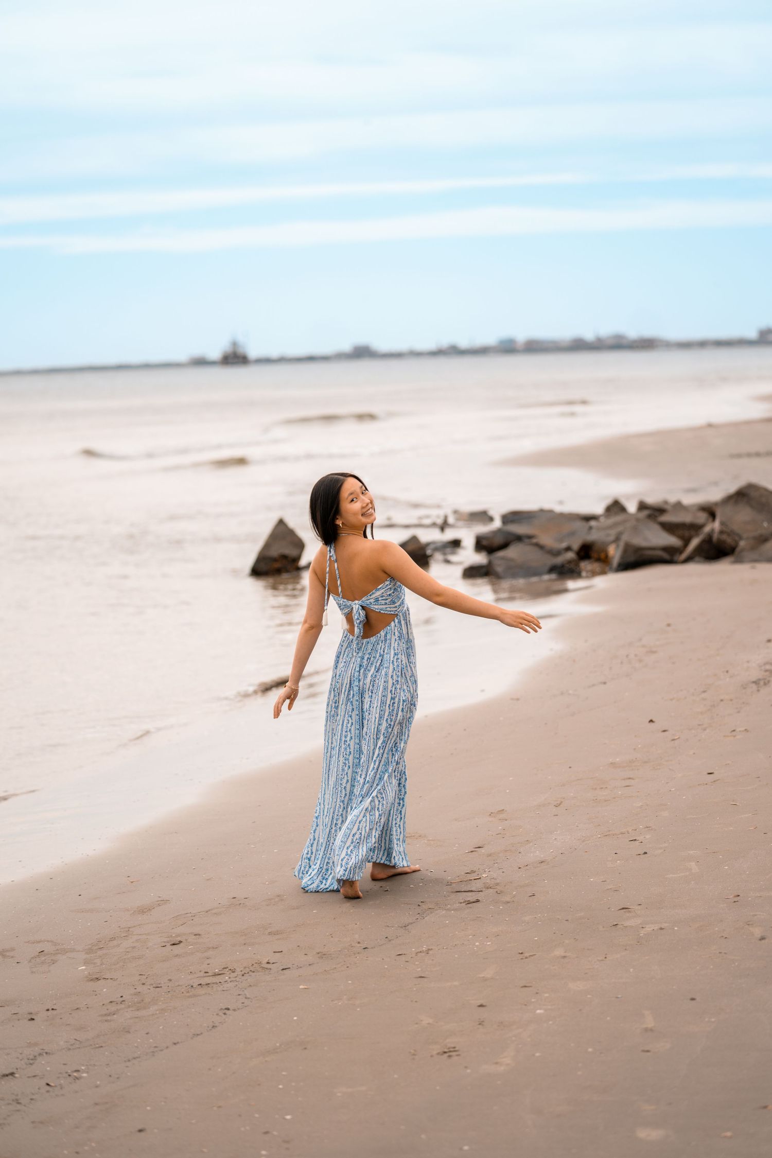 Woman in blue maxi dress with open back walks barefoot on sandy beach with arms outstretched, rocks and ocean in background.