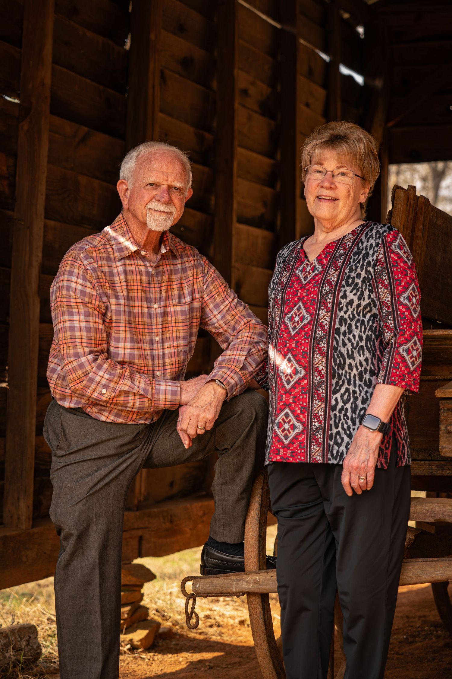 An elderly couple in casual attire poses together in a rustic barn setting with sunlight streaming in.