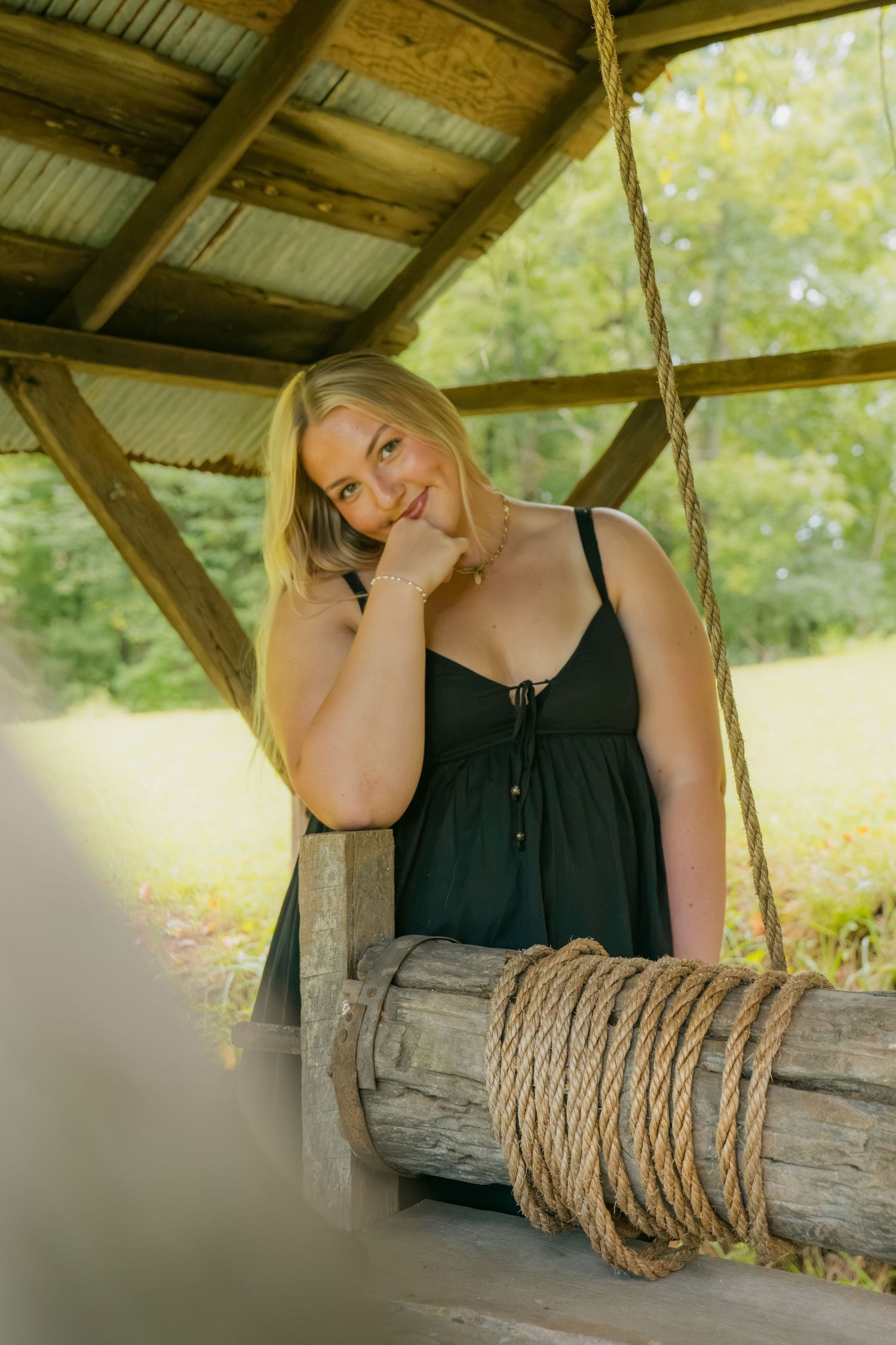 Smiling blonde woman in black dress leaning on rustic wooden swing with rope under a wooden shelter outdoors.