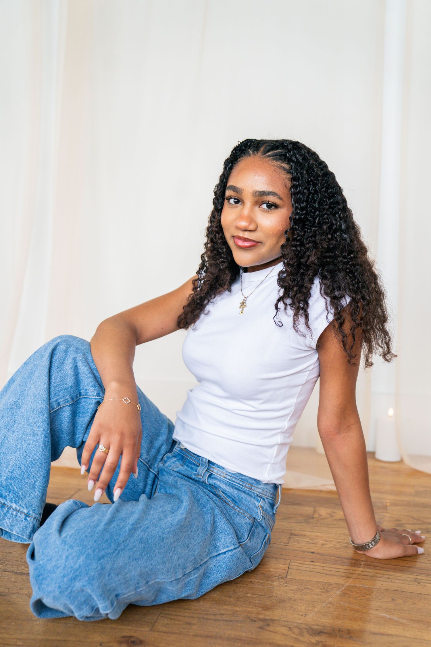 Young woman with curly hair sits on hardwood floor wearing white crop top and blue wide-leg jeans, smiling at camera.