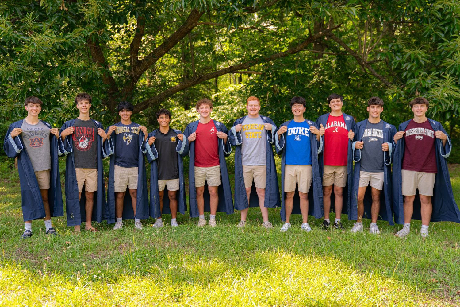 Ten young men stand in a row outdoors holding college shirts and wearing graduation capes amid lush green trees.