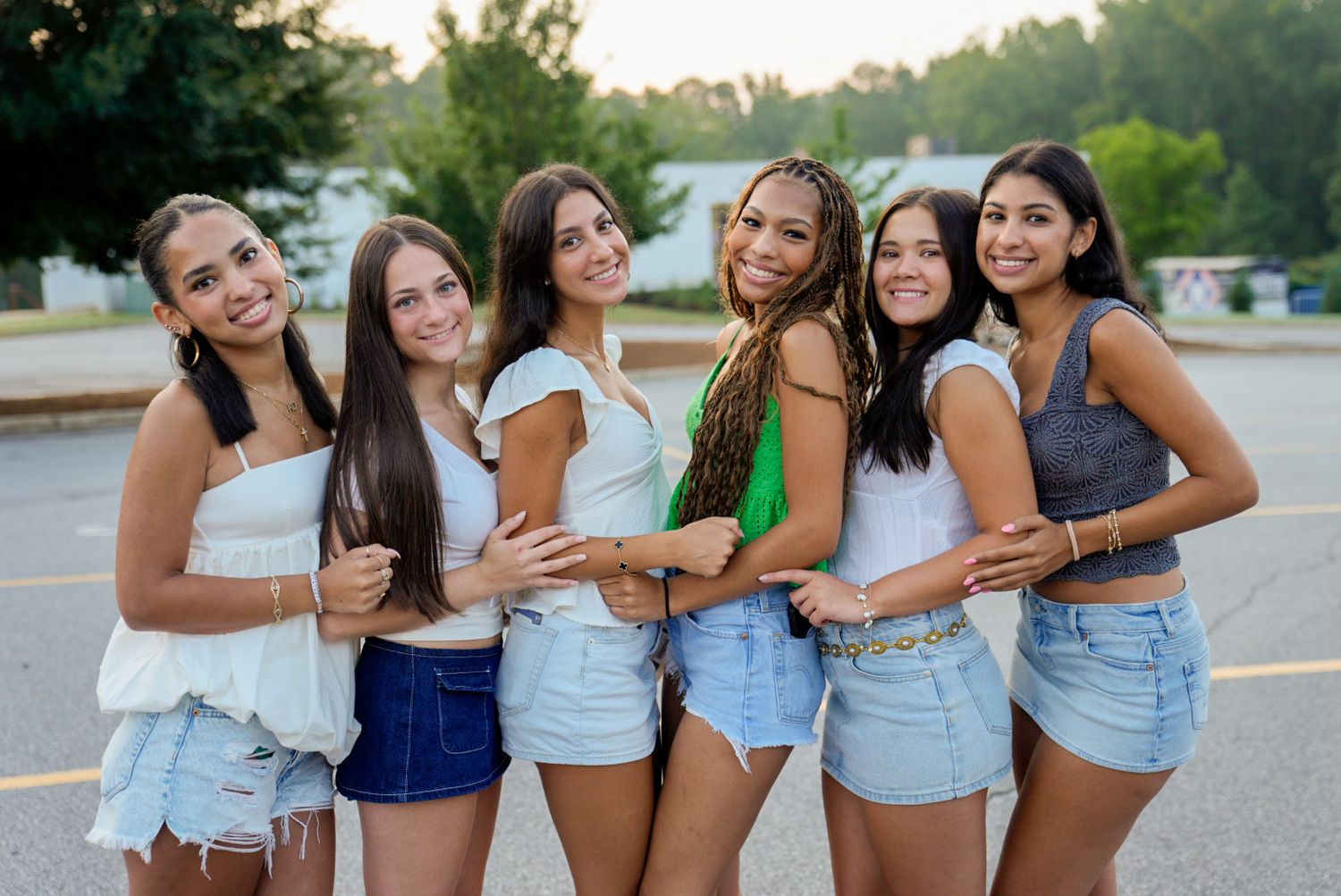 Six young women posing together outdoors in casual summer outfits, smiling and embracing in a parking lot.