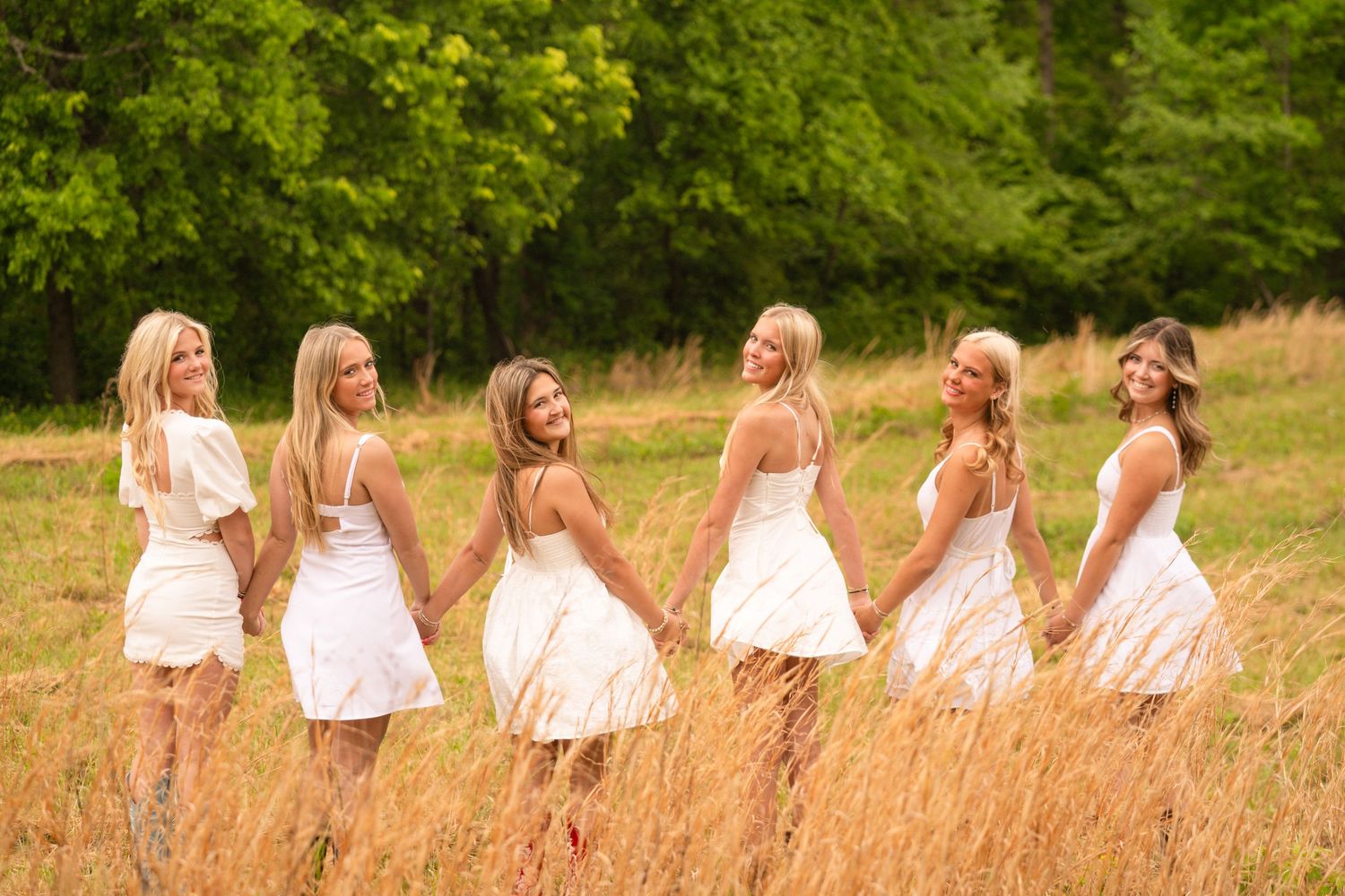 Six blonde women in white dresses hold hands in a golden field with lush green trees in the background.