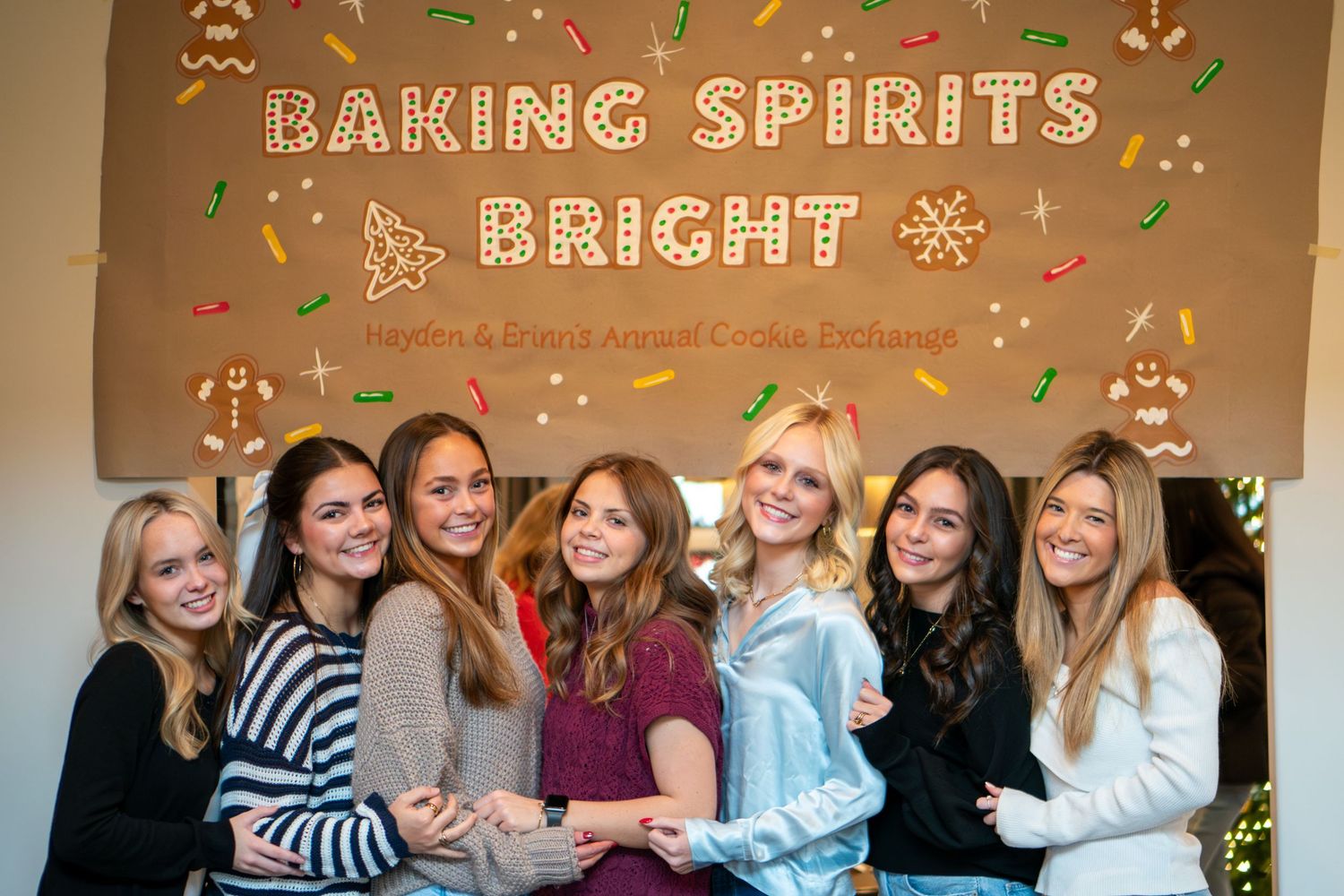 A group of friends pose together at a holiday party with decorative string lights and festive decor in the background.