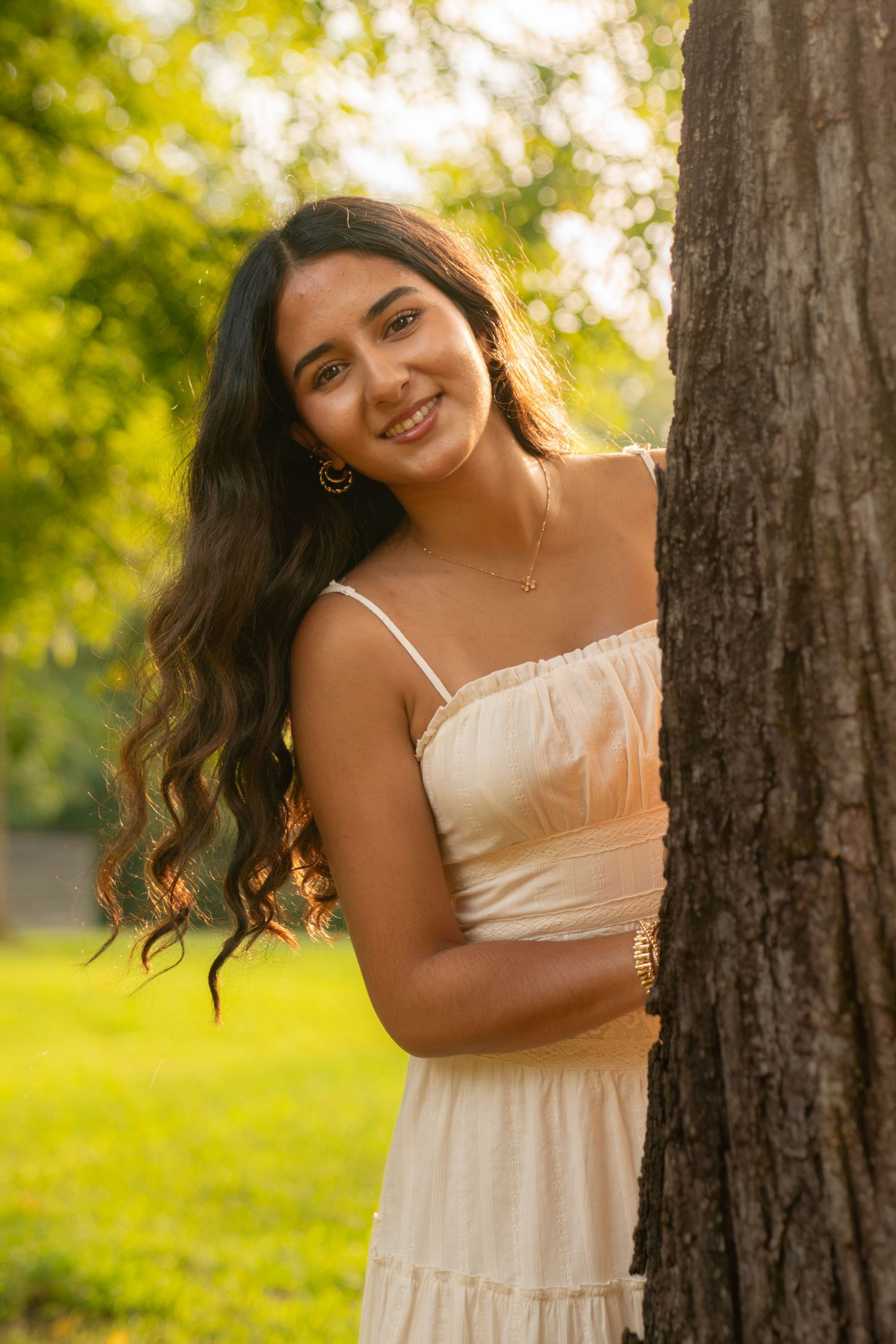 Young woman in cream sundress smiling beside a tree in a sunlit park with long wavy hair and gold jewelry.