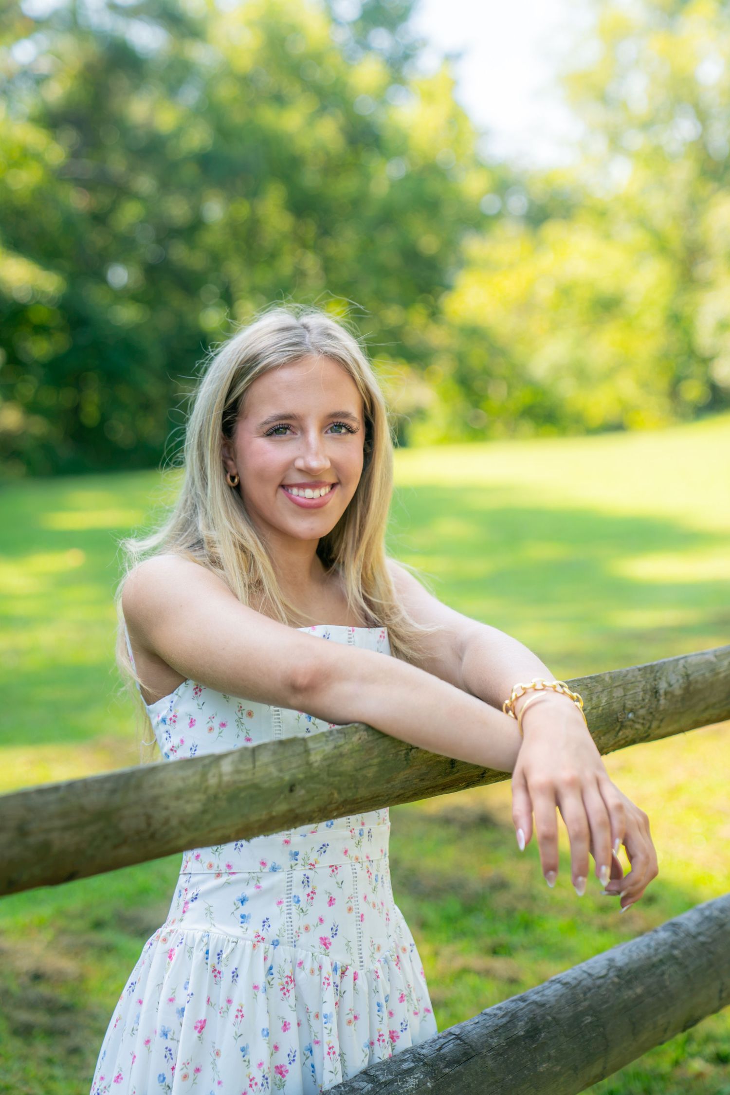 Smiling young woman with long blonde hair in a floral sundress leans on a wooden fence in a sunny, green park.