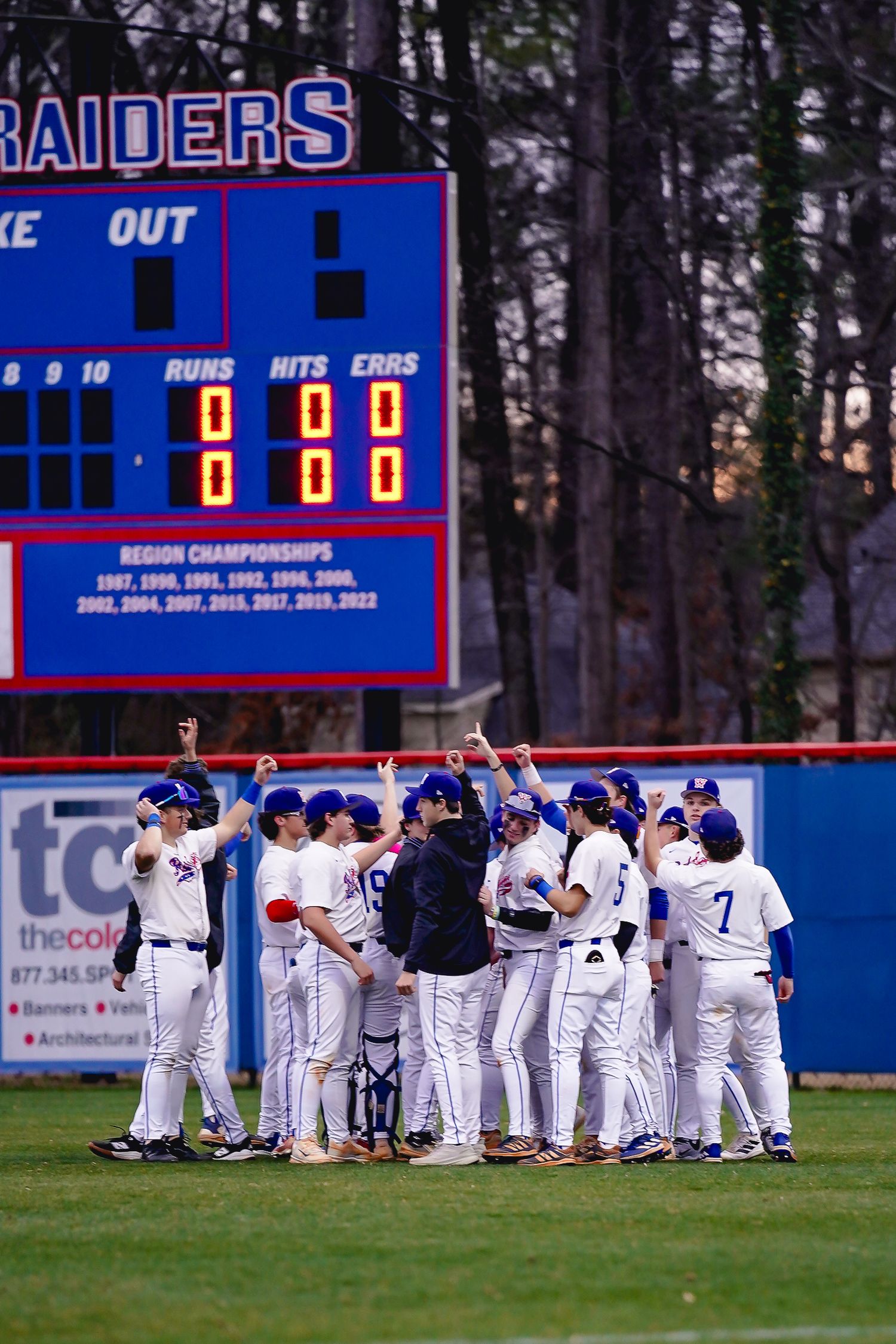 Baseball team celebrates victory on field with Raiders scoreboard visible in background.