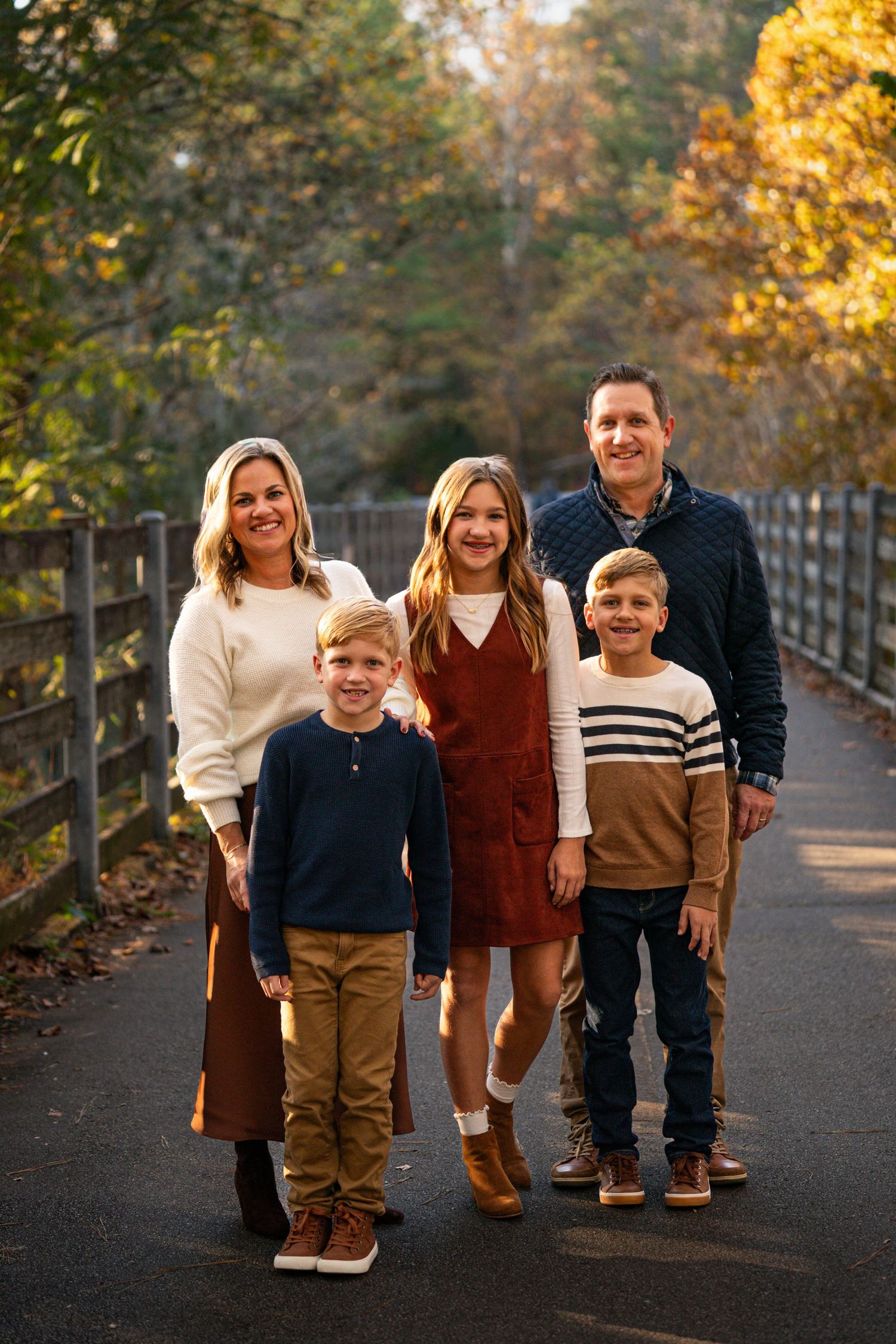 A playful family photo session on a tree-lined path during autumn with warm fall colors in the background.