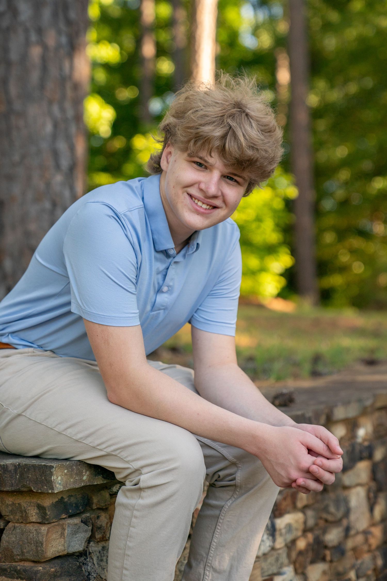 Smiling teenage boy with blonde hair wearing a light blue polo shirt, sitting on a stone wall outdoors.