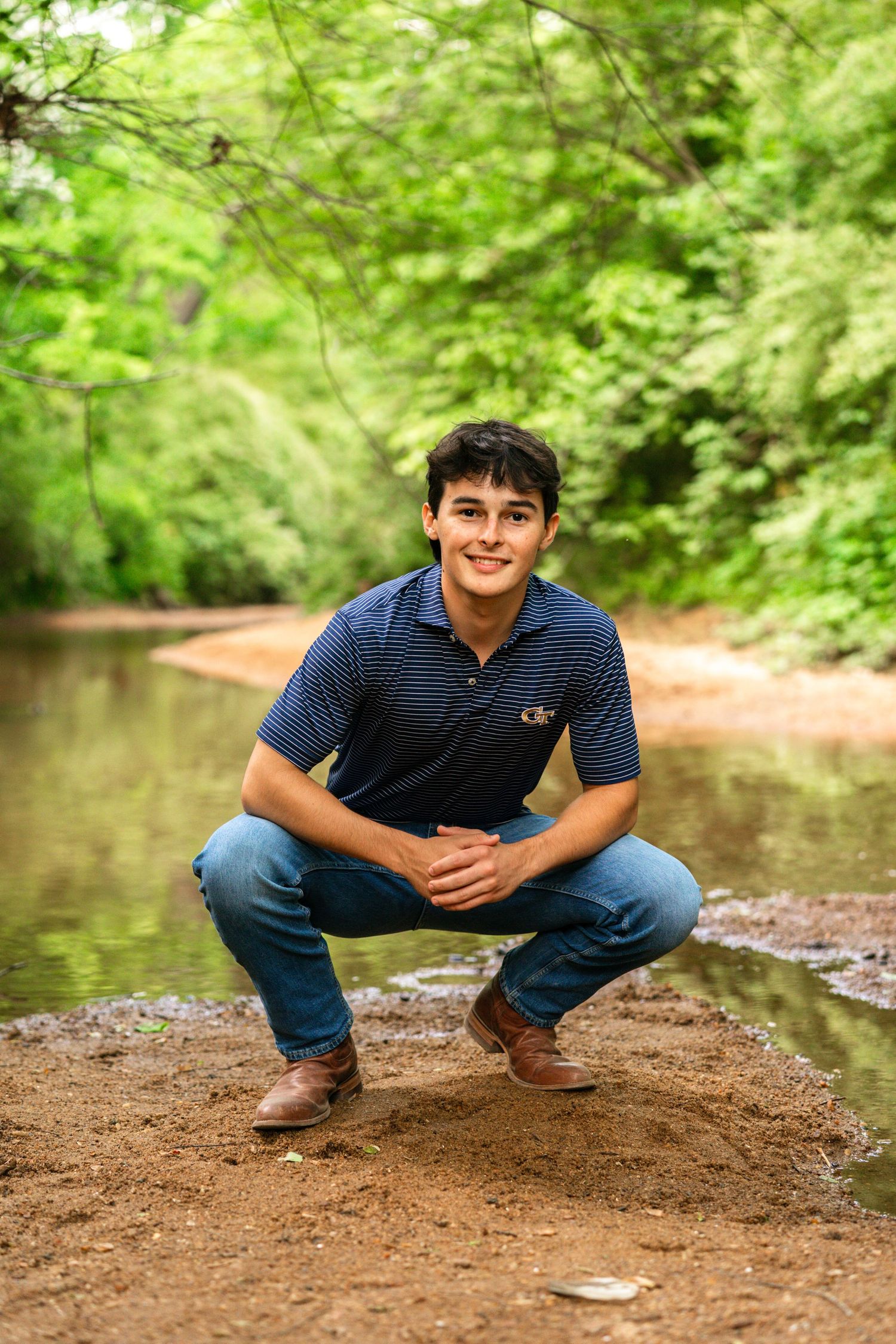 Young man in navy polo and jeans crouching by a serene creek surrounded by lush green trees.
