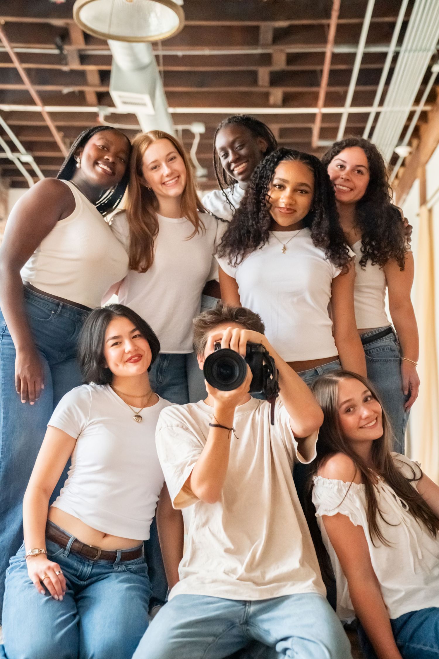 Diverse group of smiling teens in white tops and jeans pose together, one holding a camera.