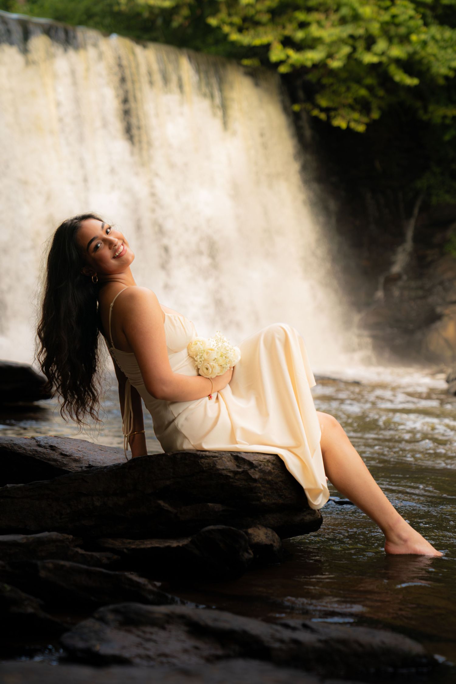 Smiling woman in cream dress holding white flowers, sitting on rocks by a cascading waterfall surrounded by lush greenery.
