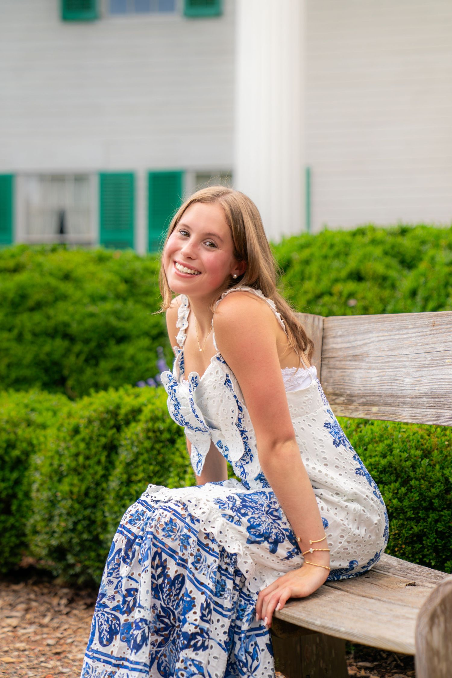 Smiling young woman in blue and white floral dress sitting on wooden bench with green bushes and white building behind her.