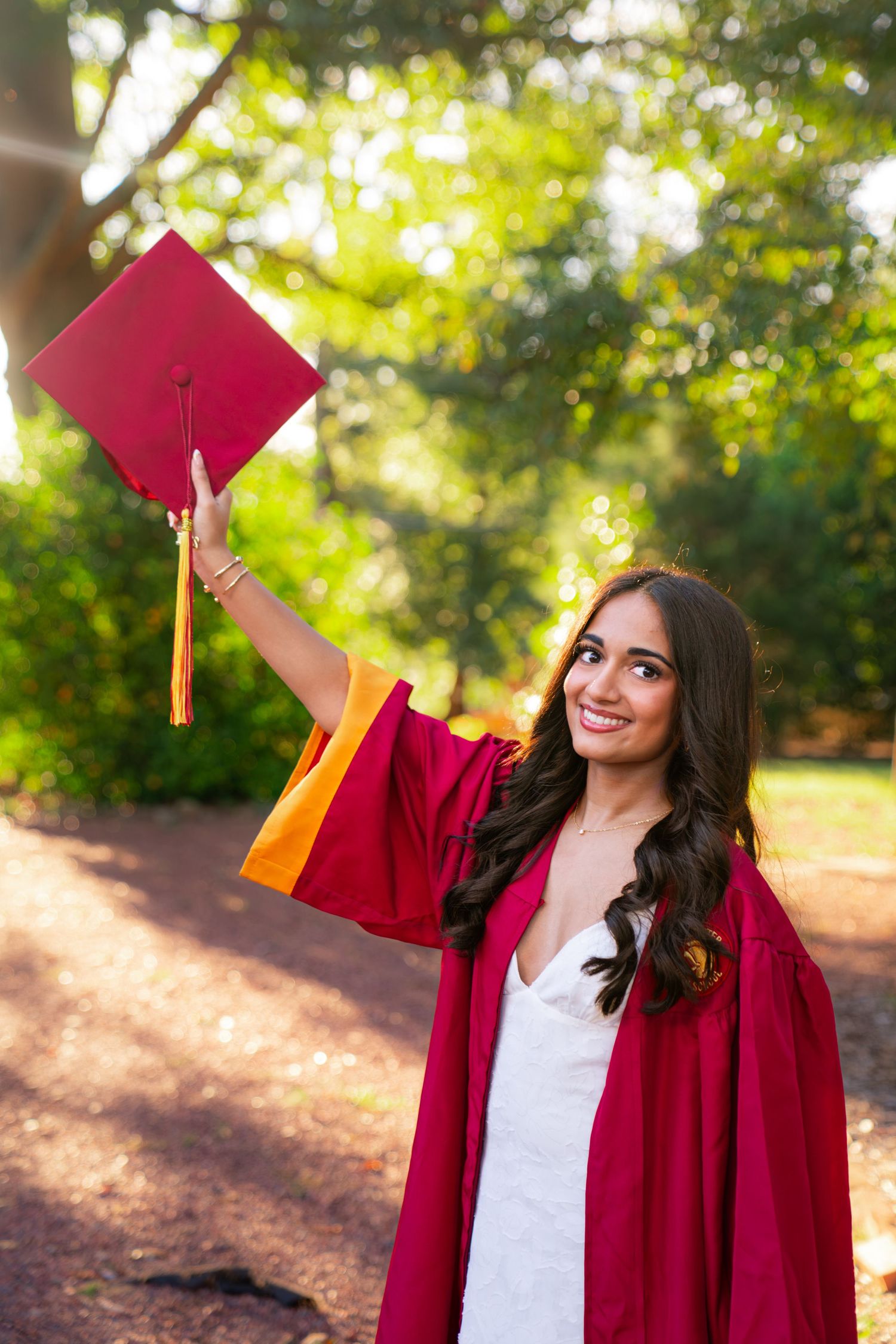 Smiling graduate in a crimson gown with gold trim holds her red mortarboard cap high while posing outdoors among sunlit green trees.