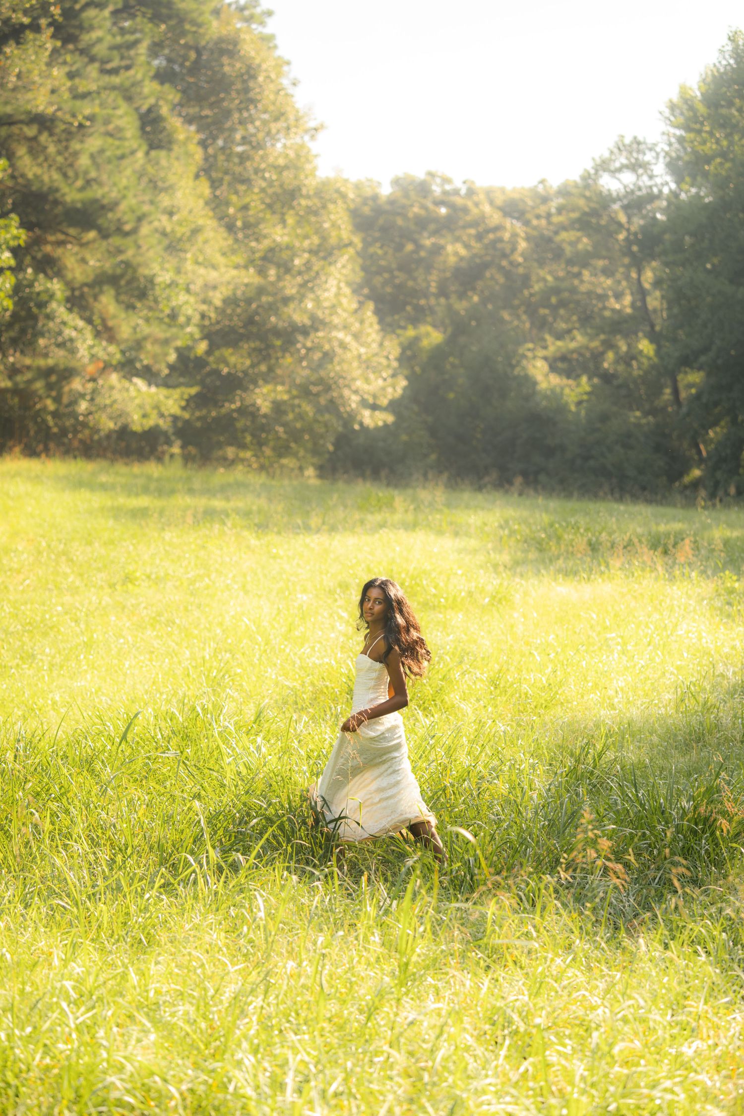 Woman in white dress walking through sunny green meadow with trees in background.
