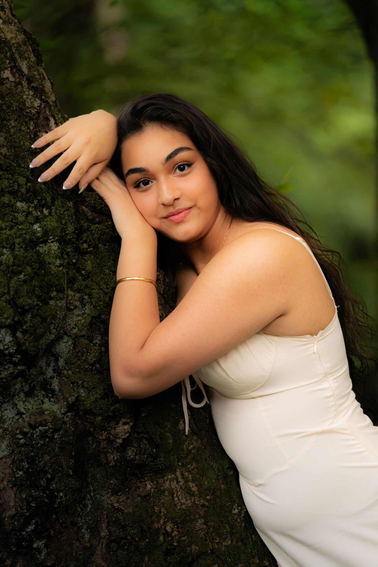 Young woman in white dress leaning against mossy tree in lush green forest, wearing gold bracelet.