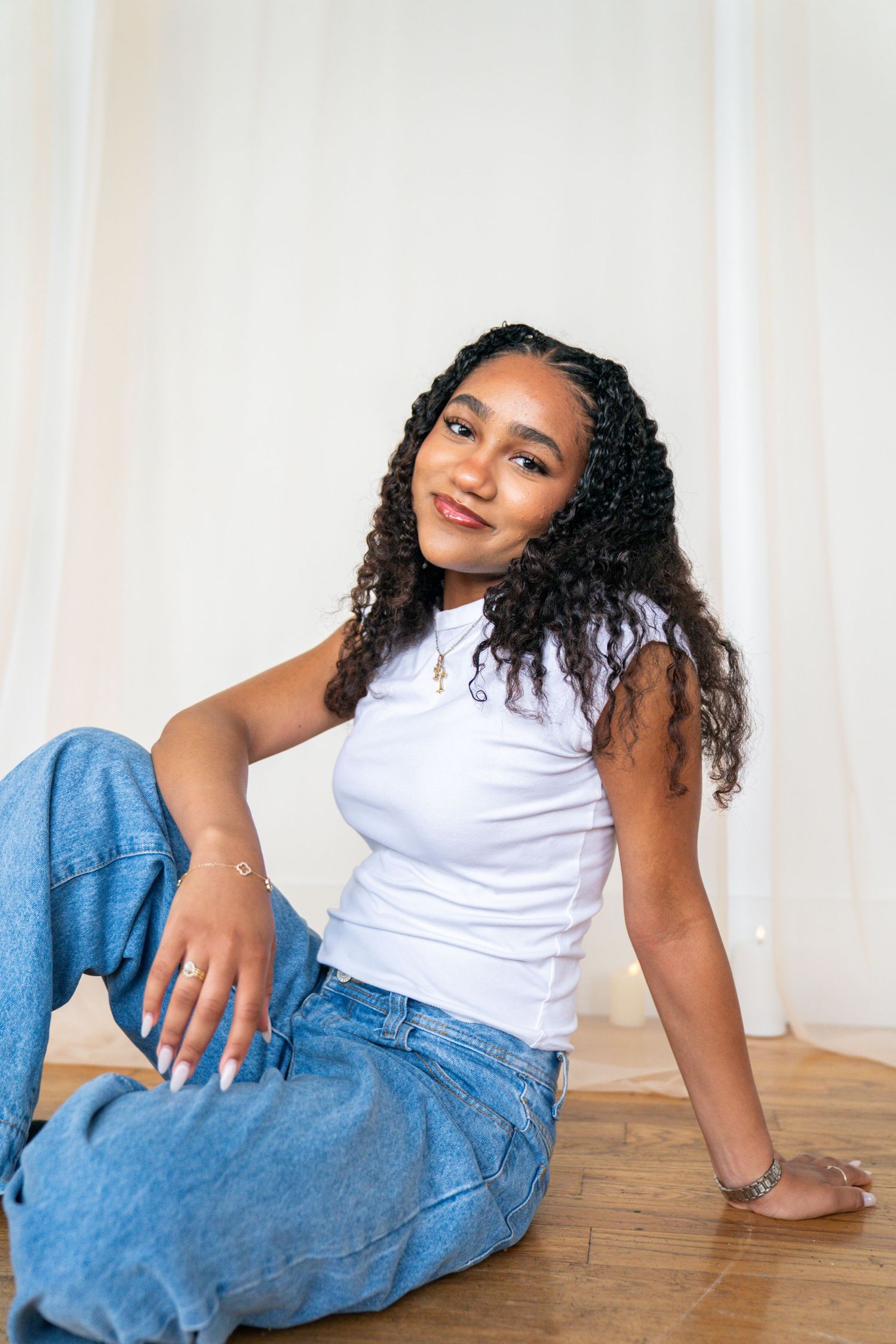 Young woman in white tank top and blue jeans sitting on floor, smiling confidently against white curtain backdrop.