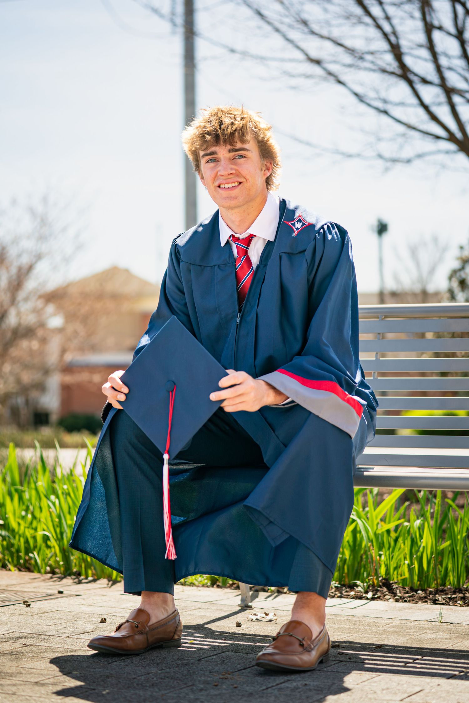 Young man in navy graduation gown and cap sitting on bench, smiling proudly on his graduation day.