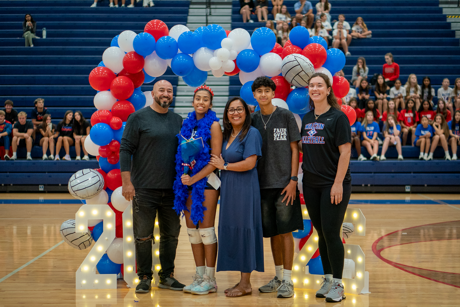 Sports team poses under red white and blue balloon arch in gymnasium.