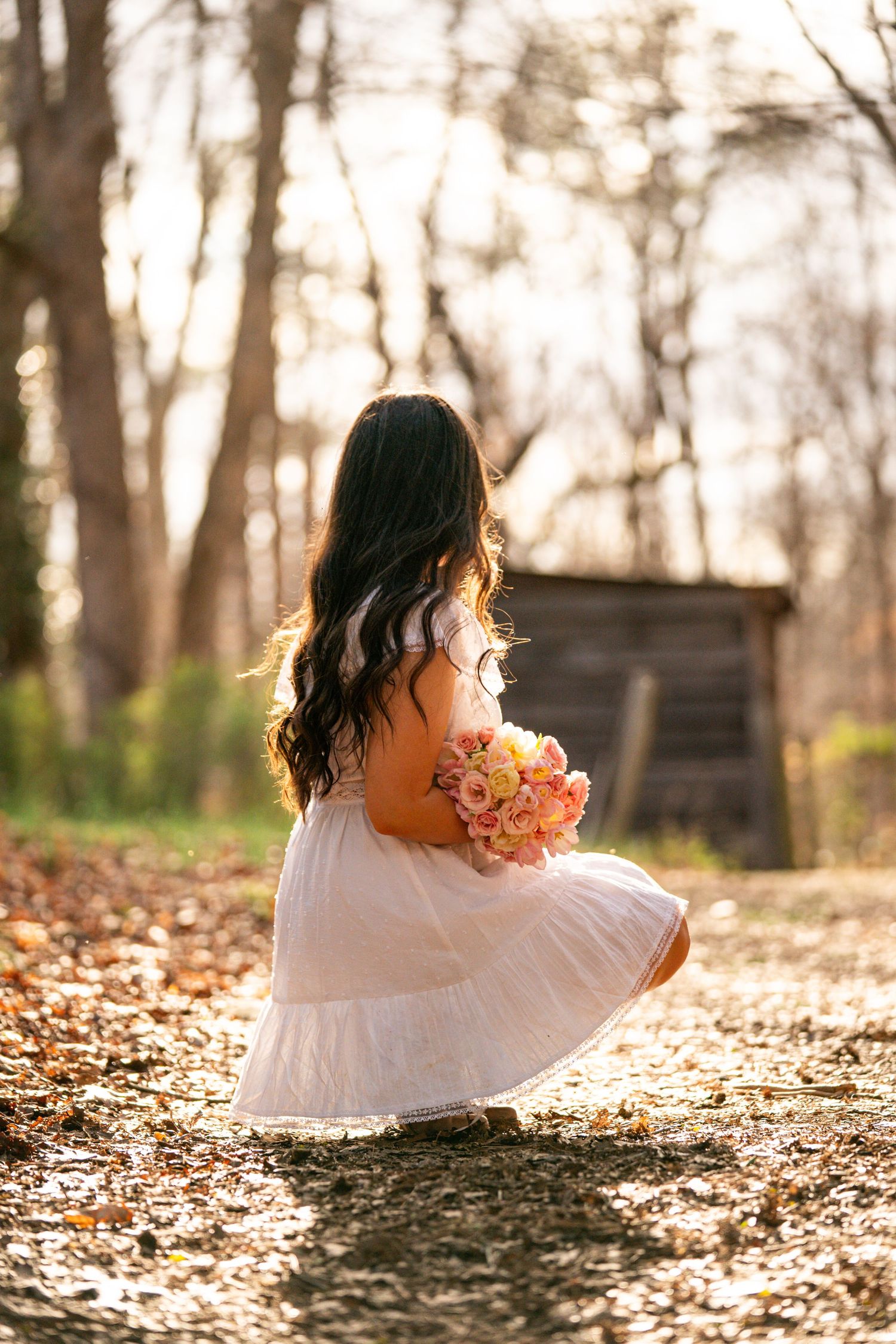 A person in a white dress sits among fallen autumn leaves during golden hour sunset in a wooded area.
