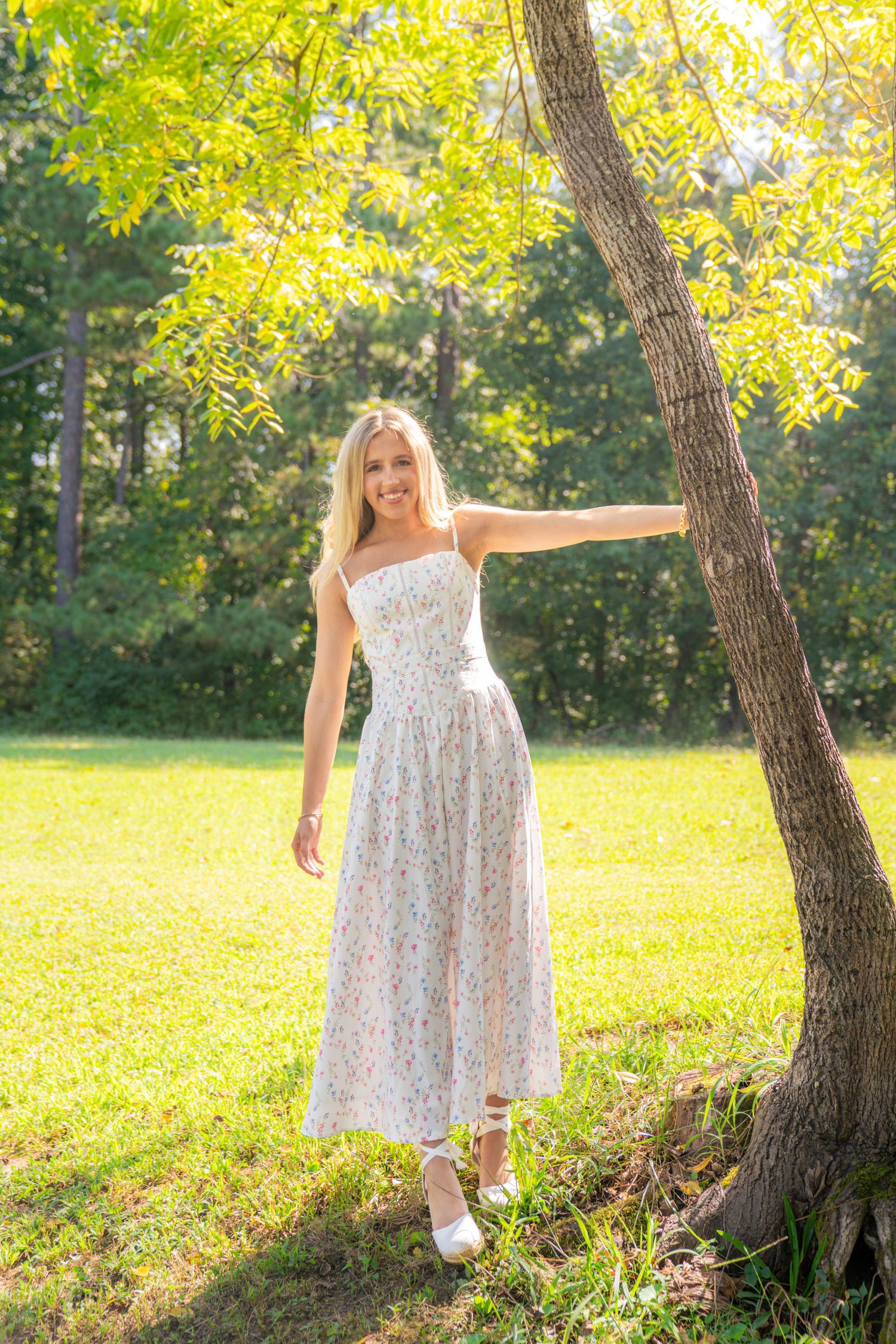 Smiling woman with long blonde hair in a white floral sundress leans on a tree trunk in a sunlit grassy park surrounded by green trees.