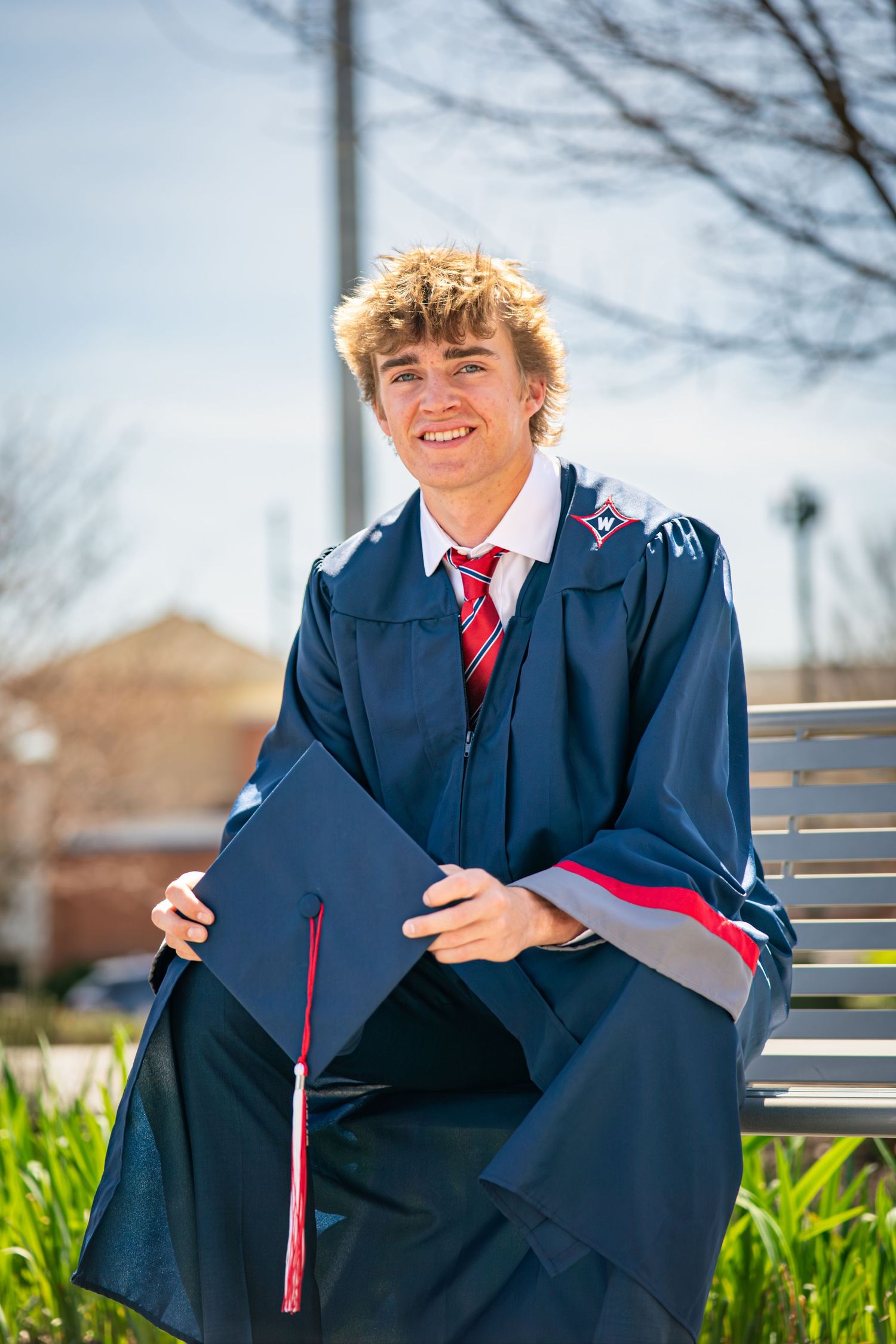 Graduate in navy blue cap and gown poses on bench outdoors during spring graduation photo session.