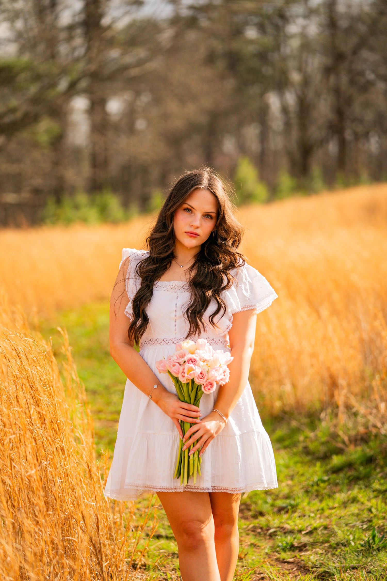 Young woman in white dress holding pink flowers bouquet standing in golden field with trees in background.
