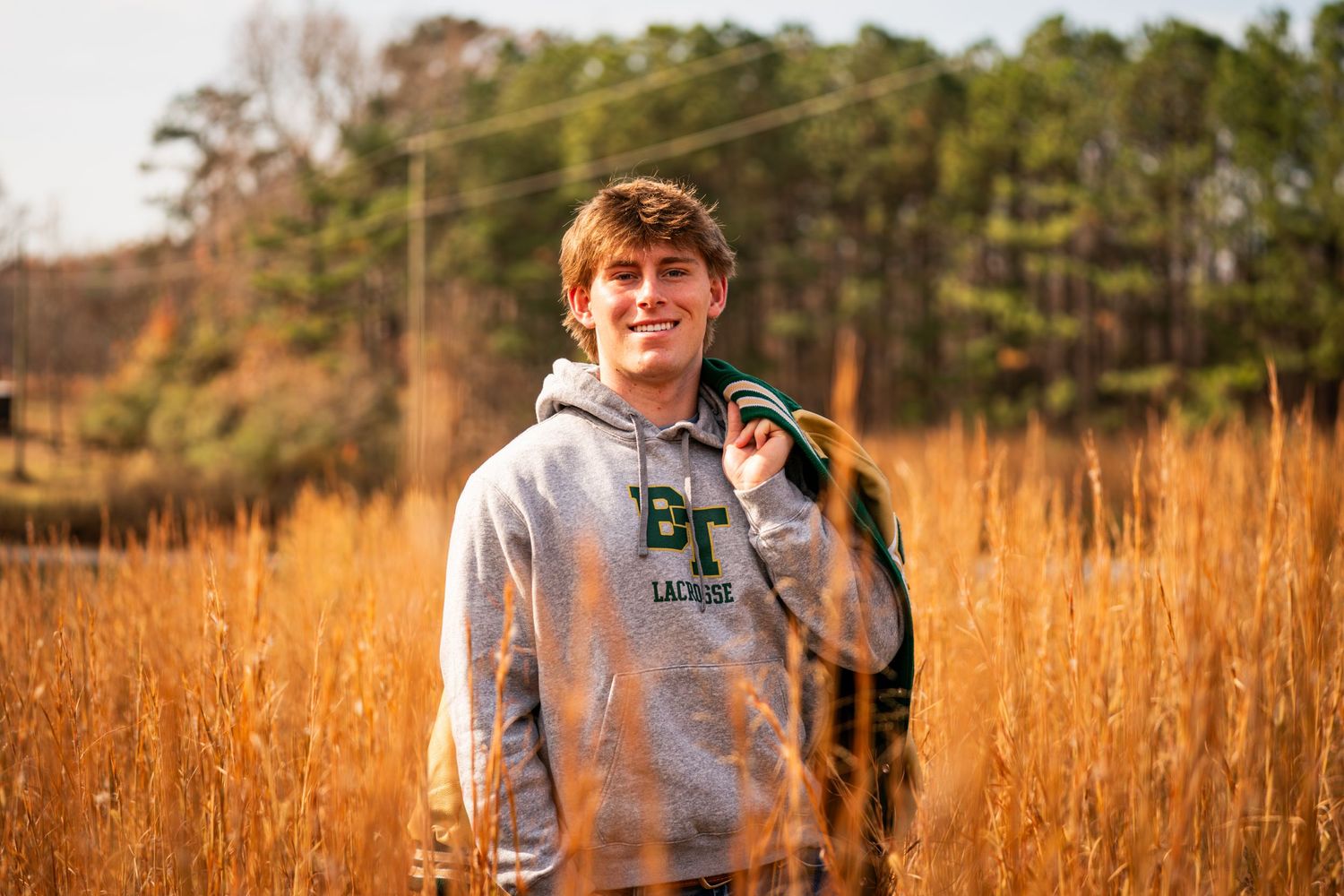 A person wearing a gray hoodie stands in a golden field of tall grass during sunset with pine trees in the background.
