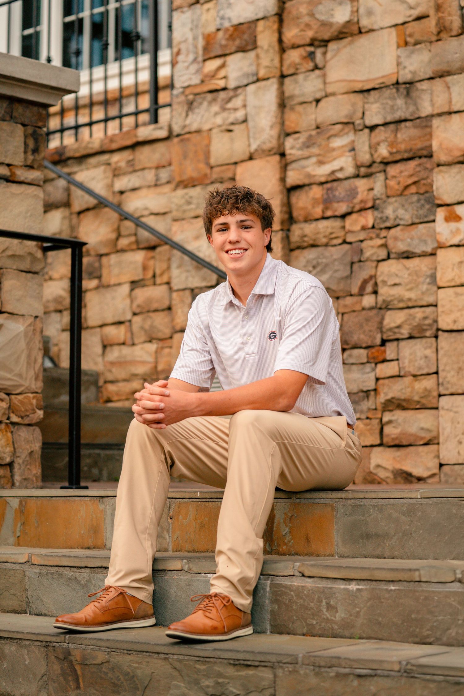 Young man in khaki pants and white polo sits smiling on stone steps outside a brick building.