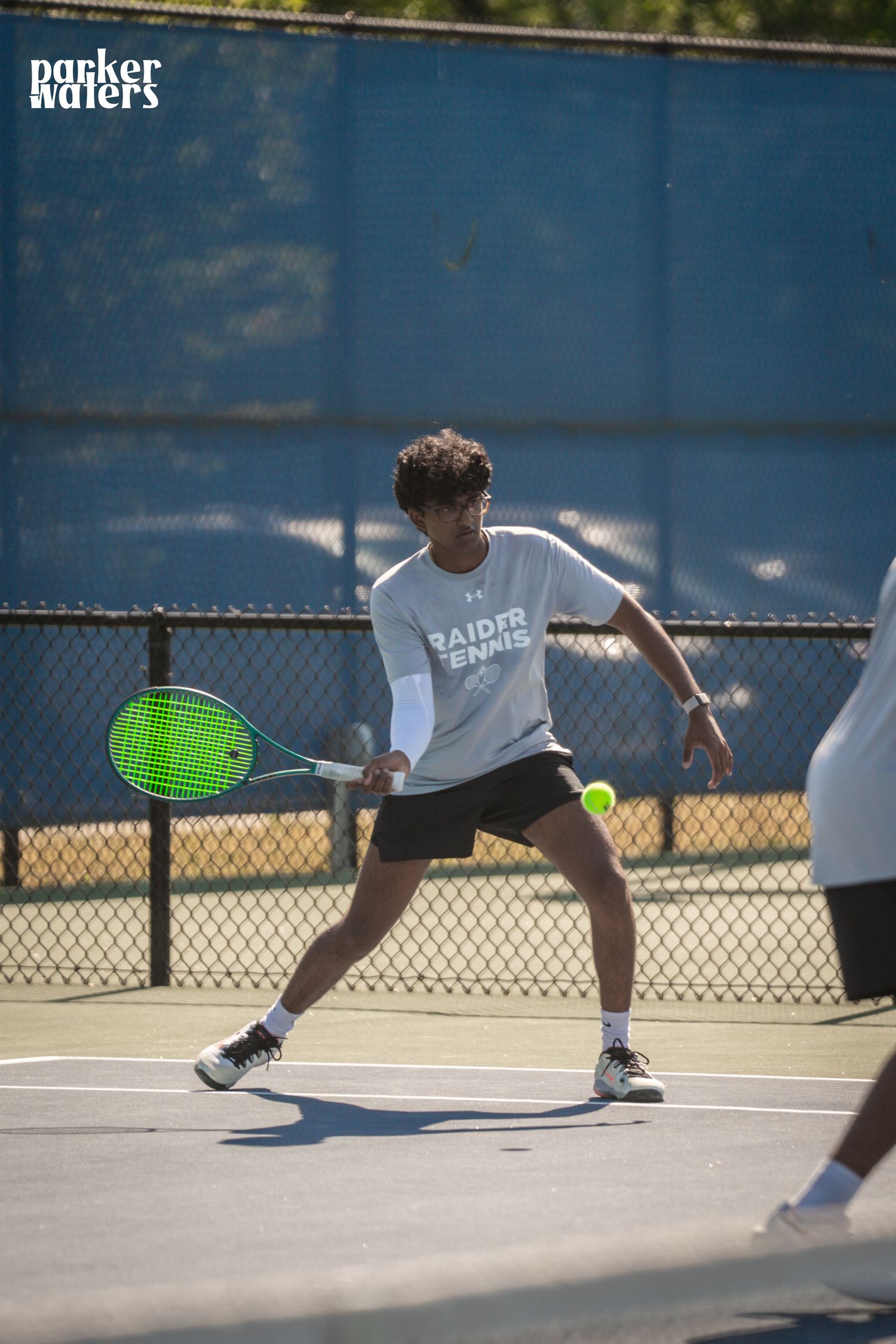 A tennis player in a Raiders Tennis shirt hits a forehand shot on an outdoor court in bright sunlight.