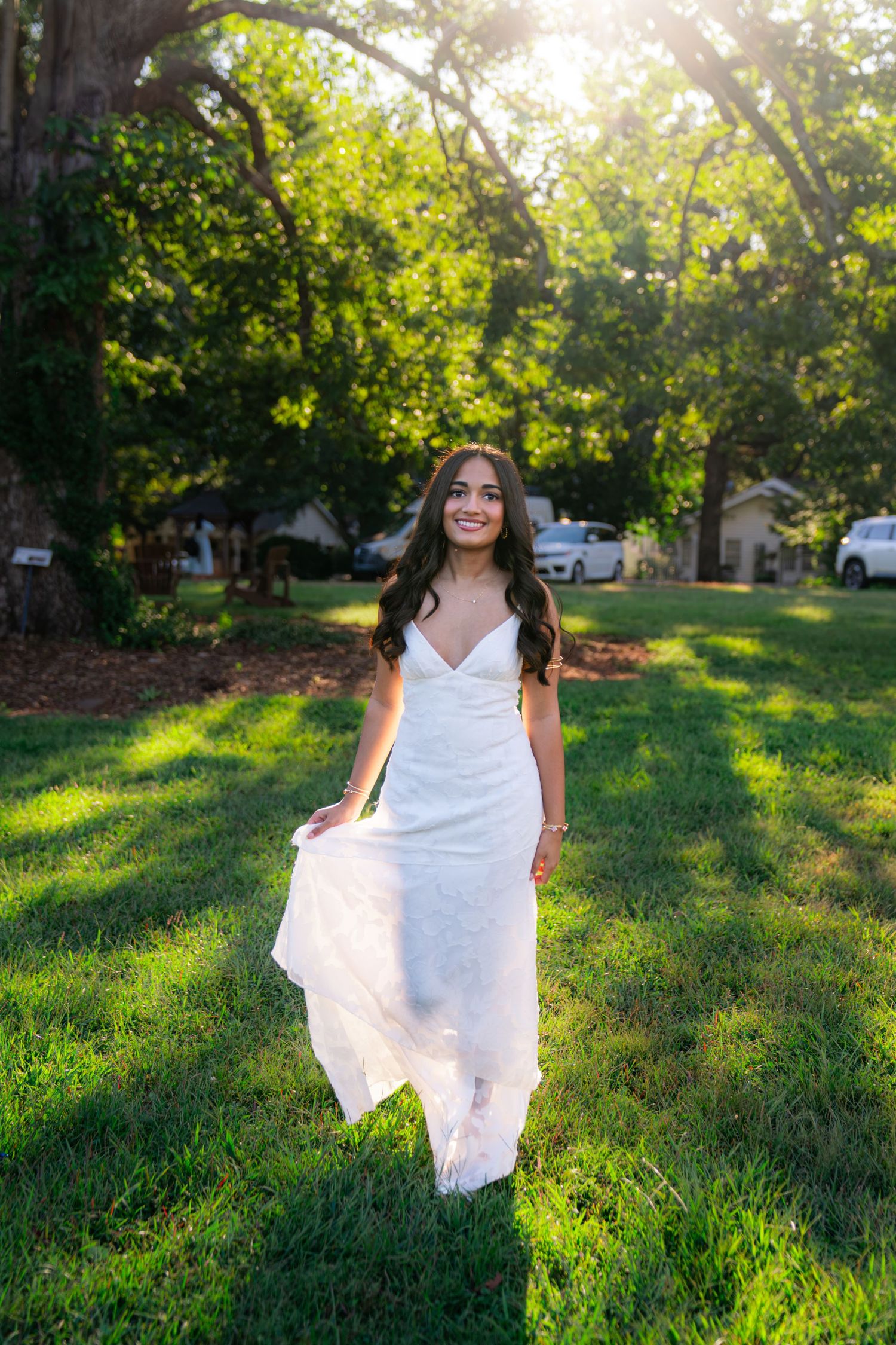 Young woman with long dark hair in a white spaghetti-strap dress walks across a sunlit green lawn beneath large shady trees.