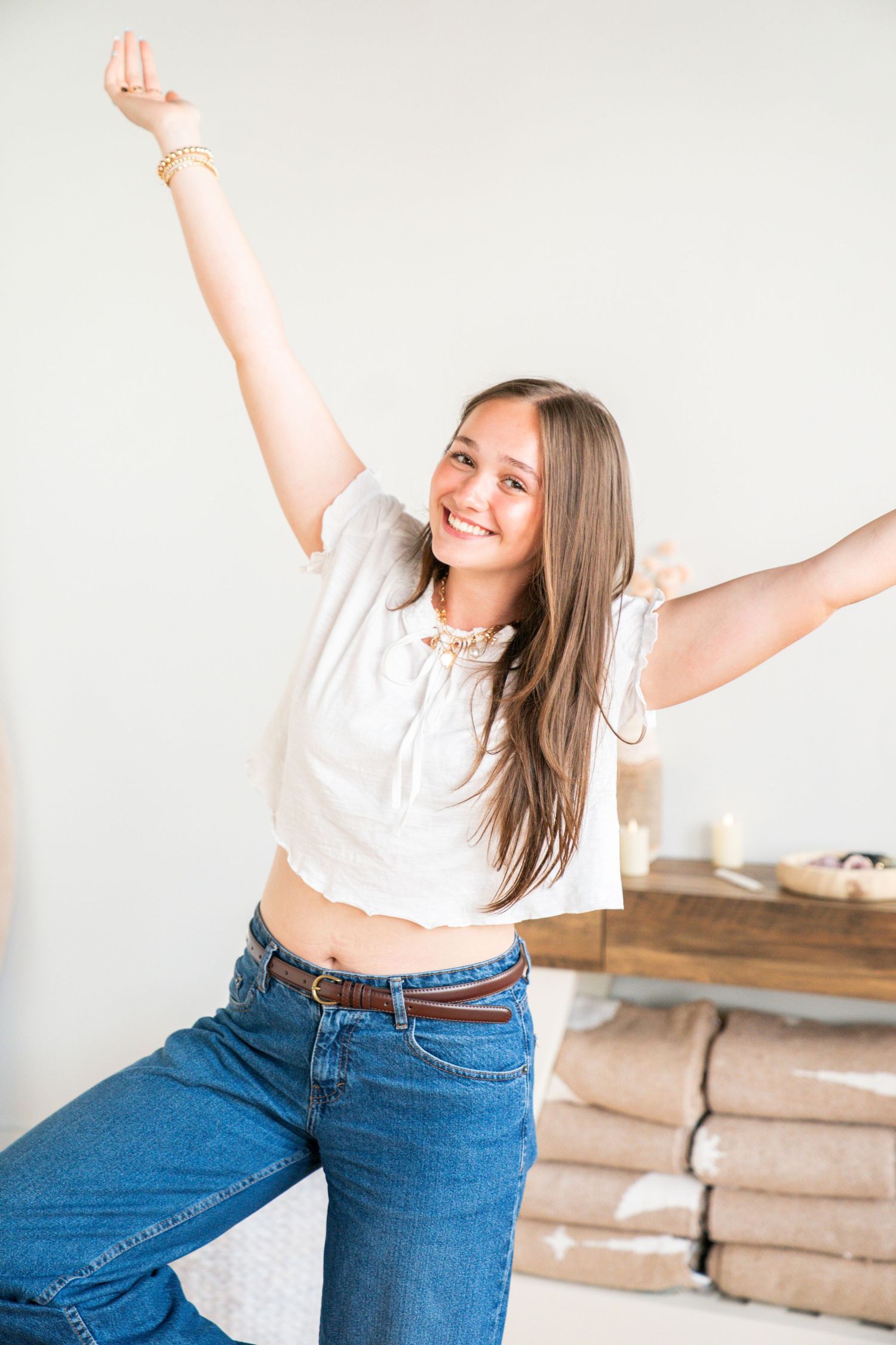 Happy young woman in white crop top and jeans stretching arms wide open, smiling joyfully indoors.