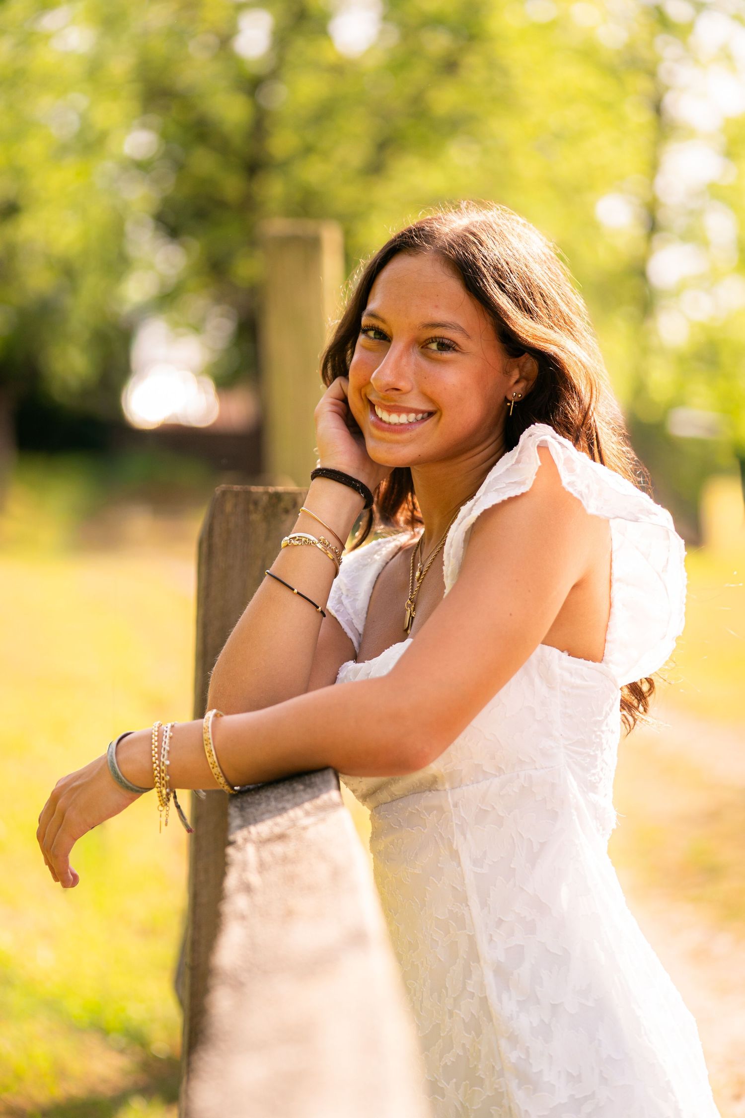 Young woman in white dress smiling outdoors, leaning on wooden fence post in sunny park setting.