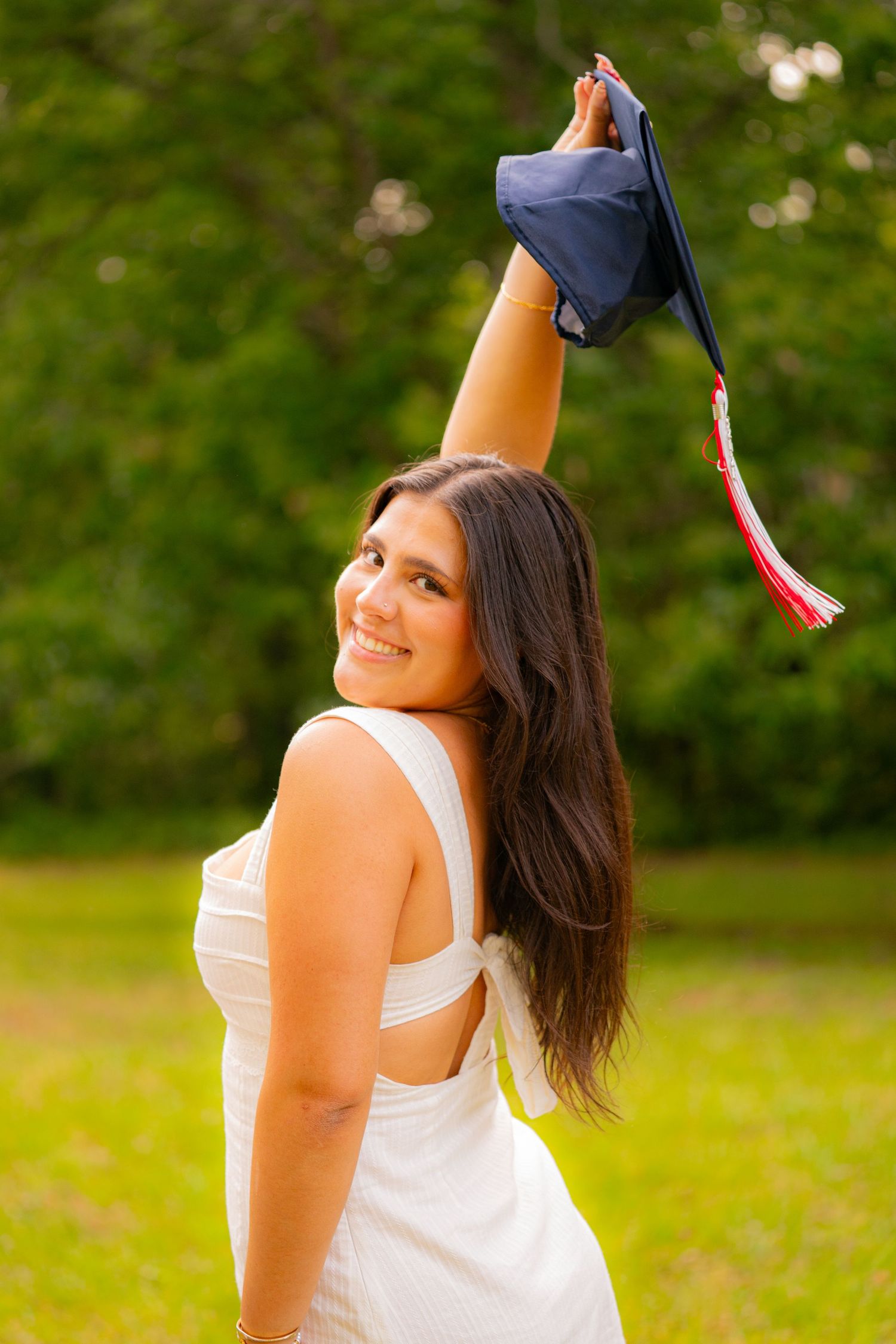 Smiling young woman in white dress holding graduation cap aloft outdoors, celebrating academic achievement.