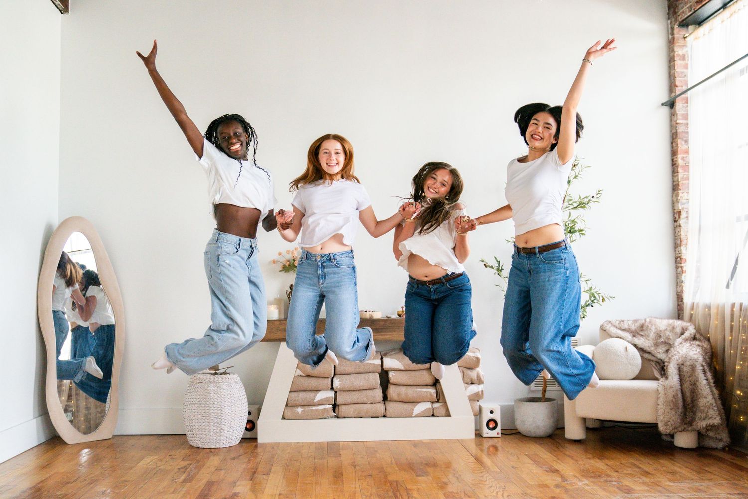 Four diverse women jumping joyfully in a bright modern room with hardwood floors and white walls.