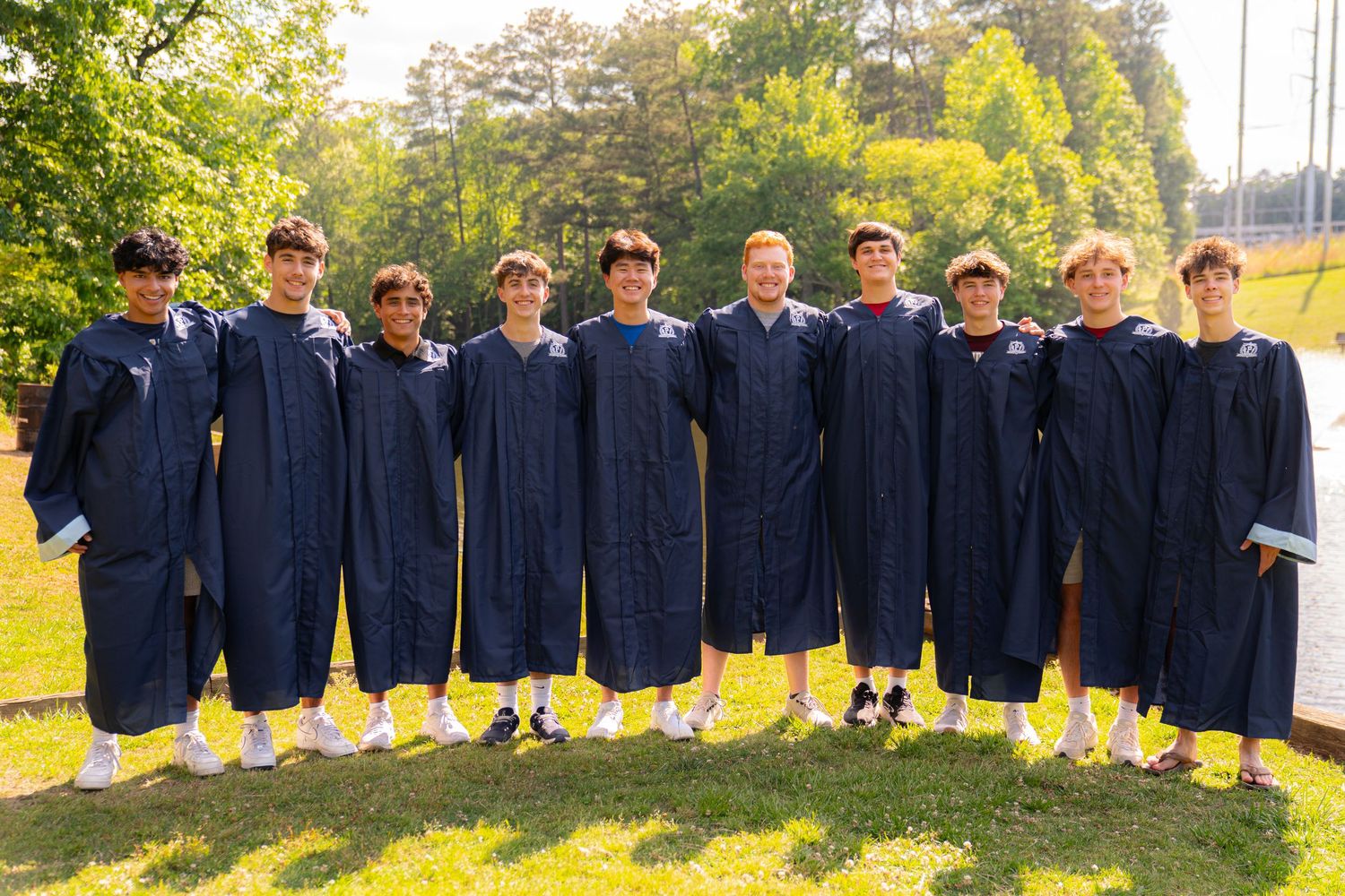 Group of ten students in navy blue graduation gowns posing outdoors on green grass with trees in background.