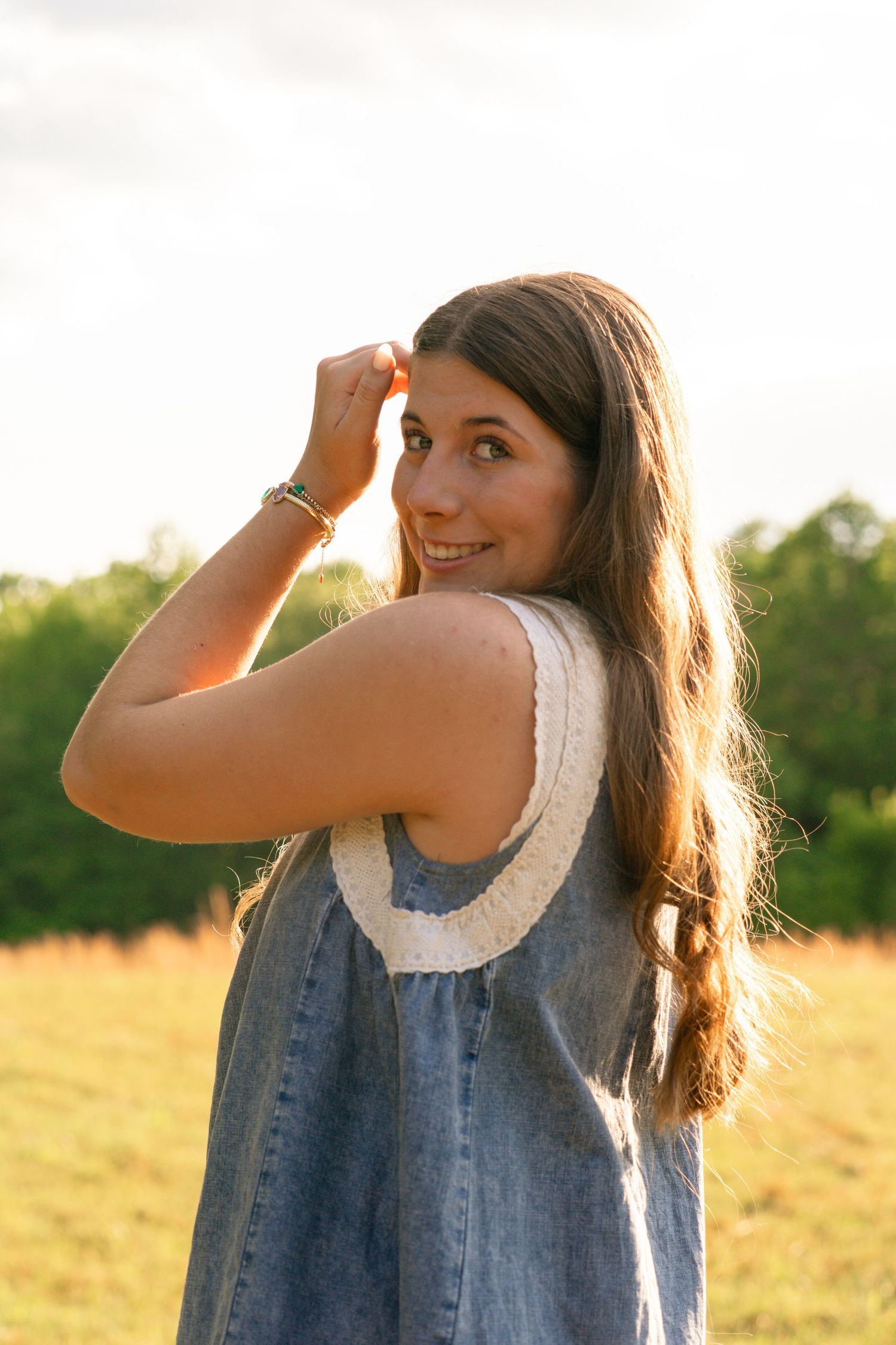 Young woman in denim dress smiling over shoulder in golden hour sunlight with green field background.