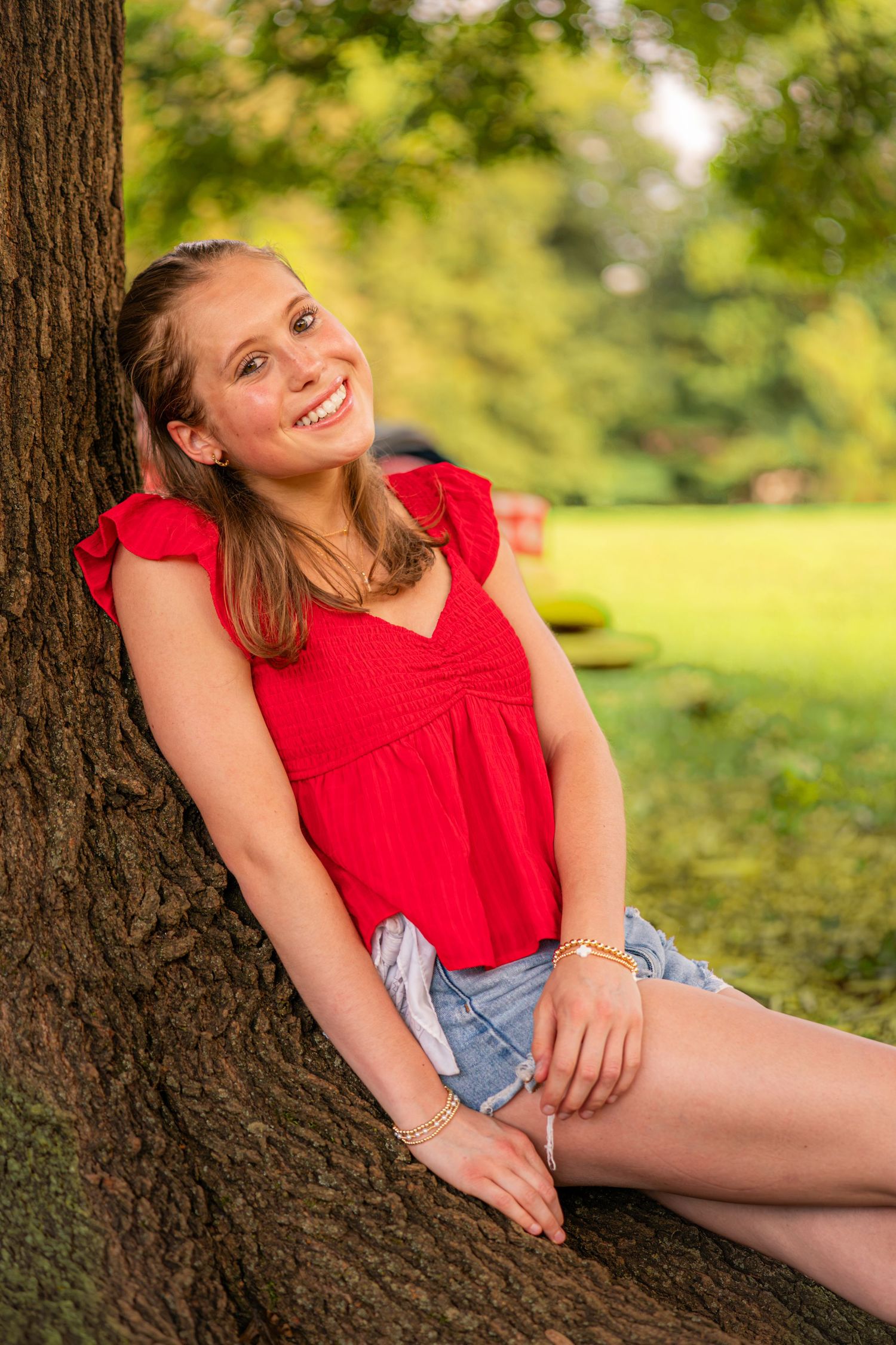Young woman in red top and denim shorts smiling while sitting against a tree in a sunny green park.