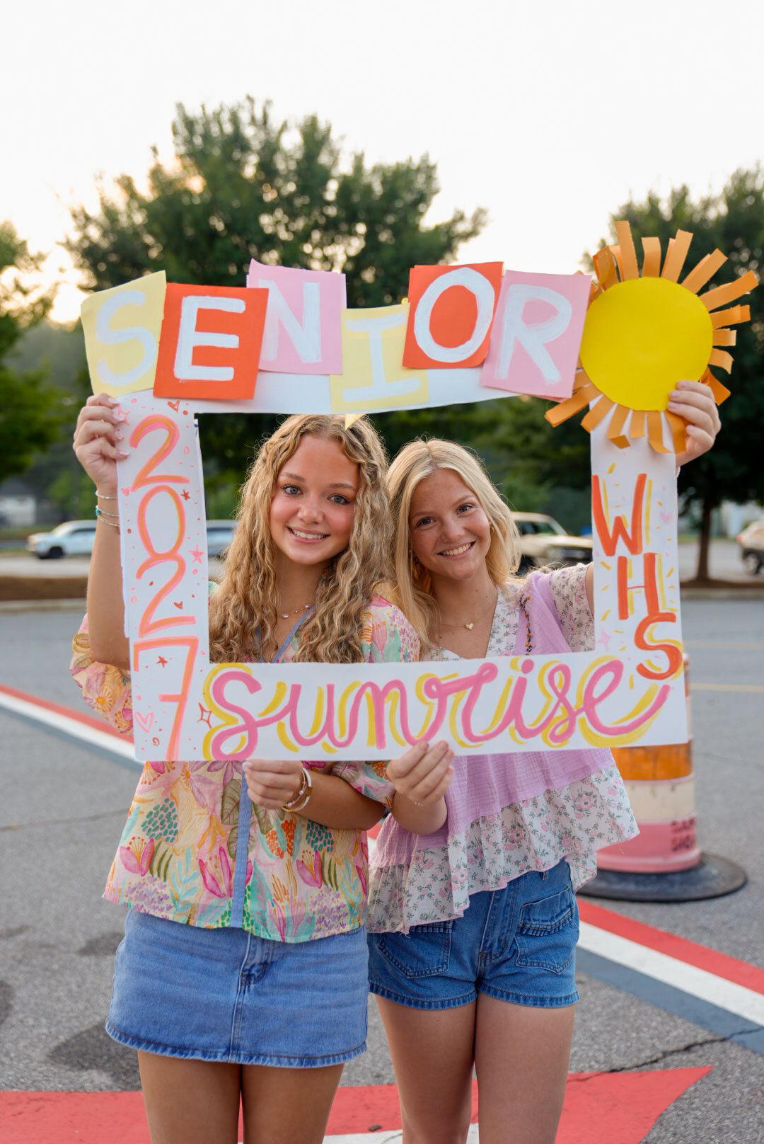 Two blonde teen girls hold colorful handmade Senior 2027 Sunrise signs in a parking lot at golden hour.