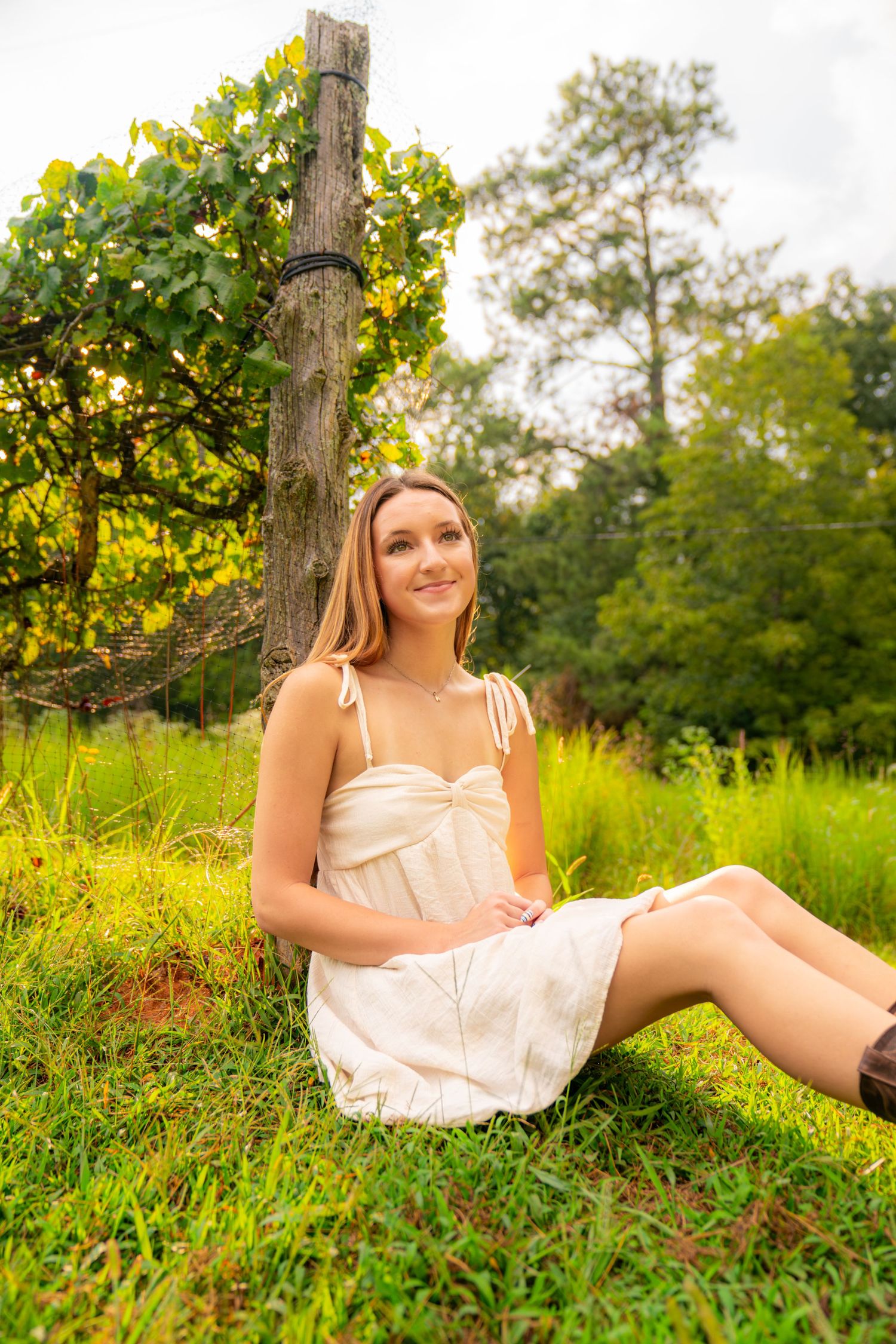 Young woman in white sundress sitting on grass near wooden post surrounded by lush green trees outdoors.