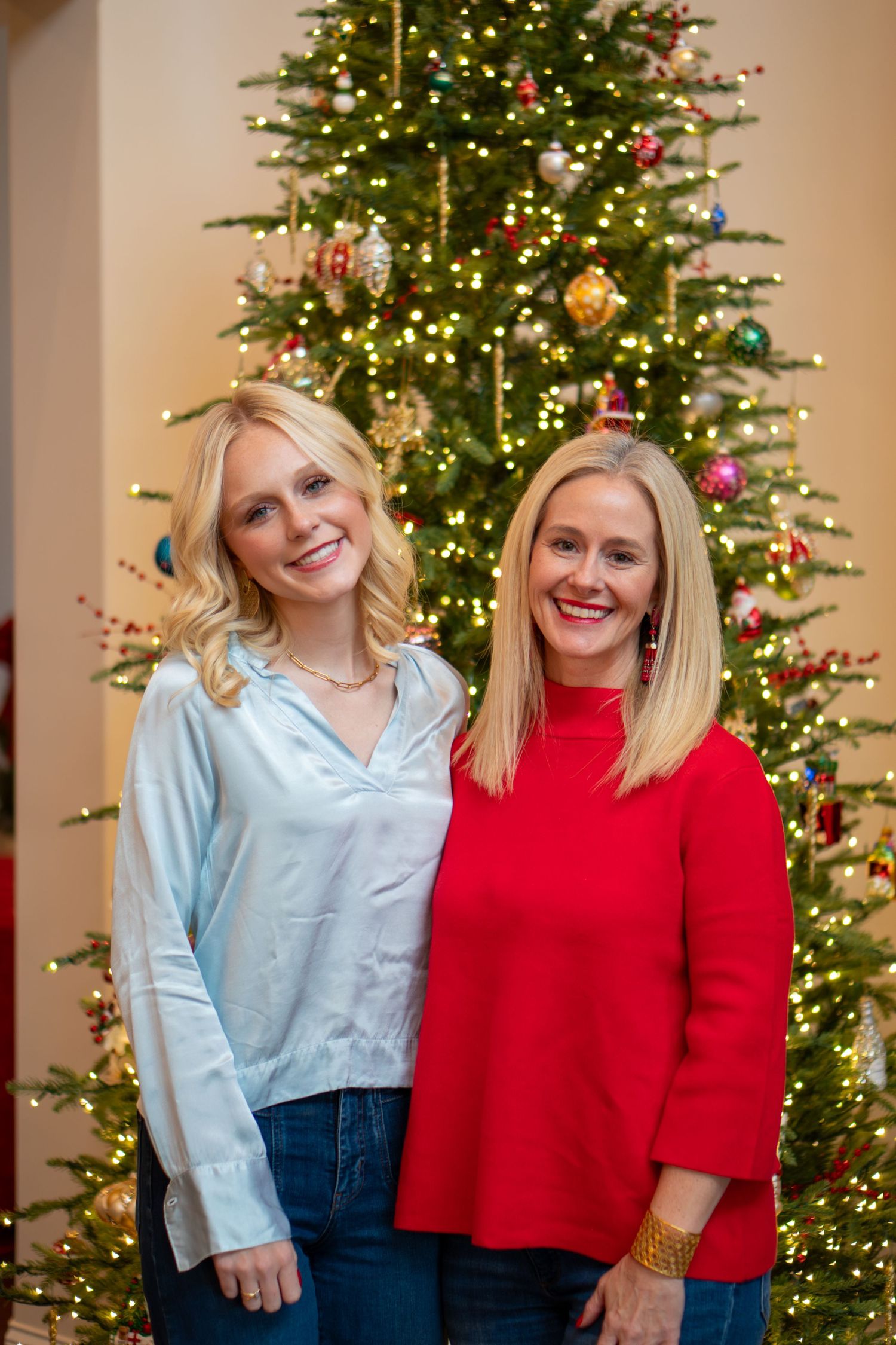 Two people wearing festive holiday outfits pose together in front of a decorated Christmas tree with twinkling lights.