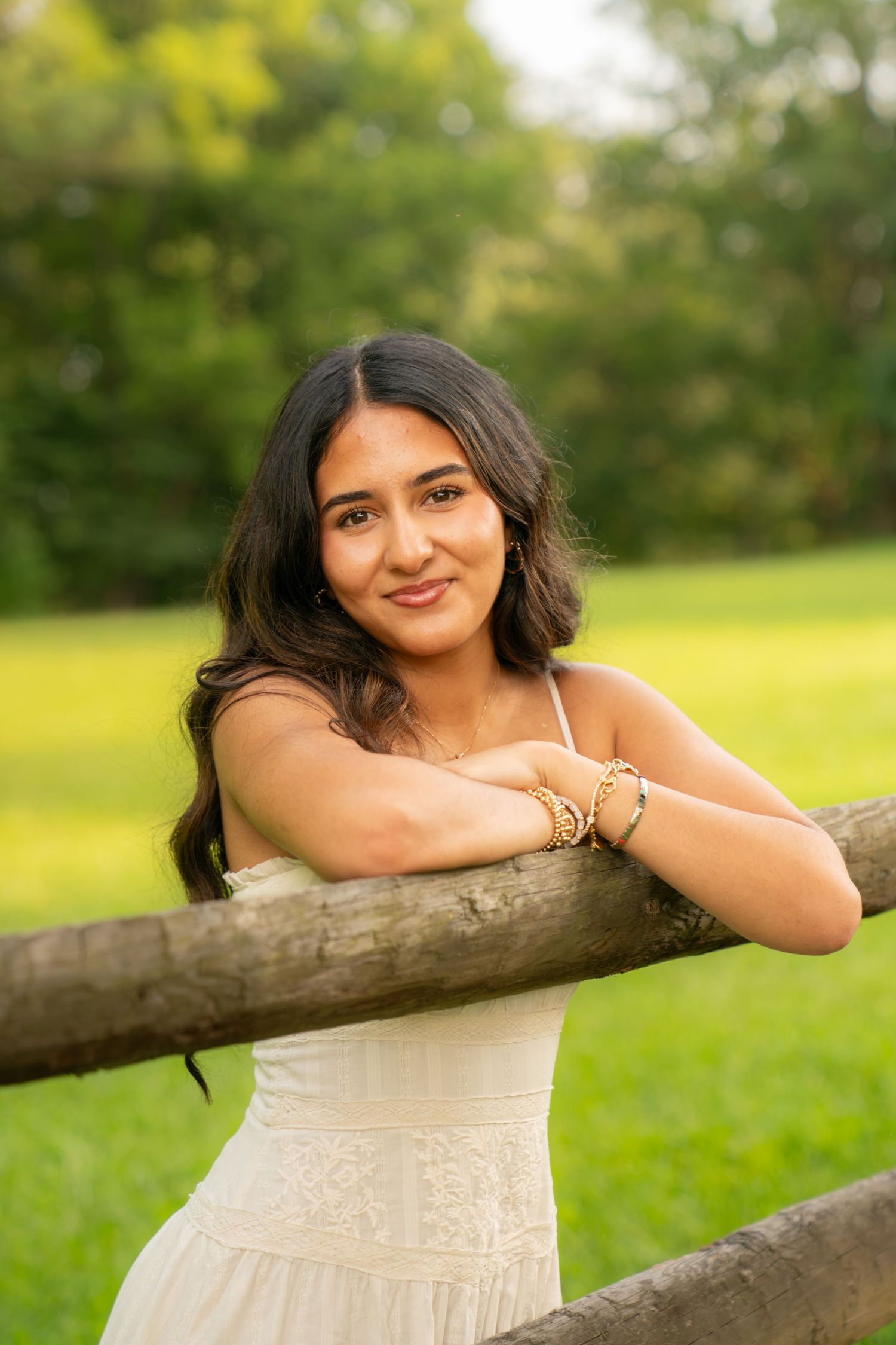 Young woman in white dress leaning on wooden fence rail in lush green park, smiling warmly at camera.