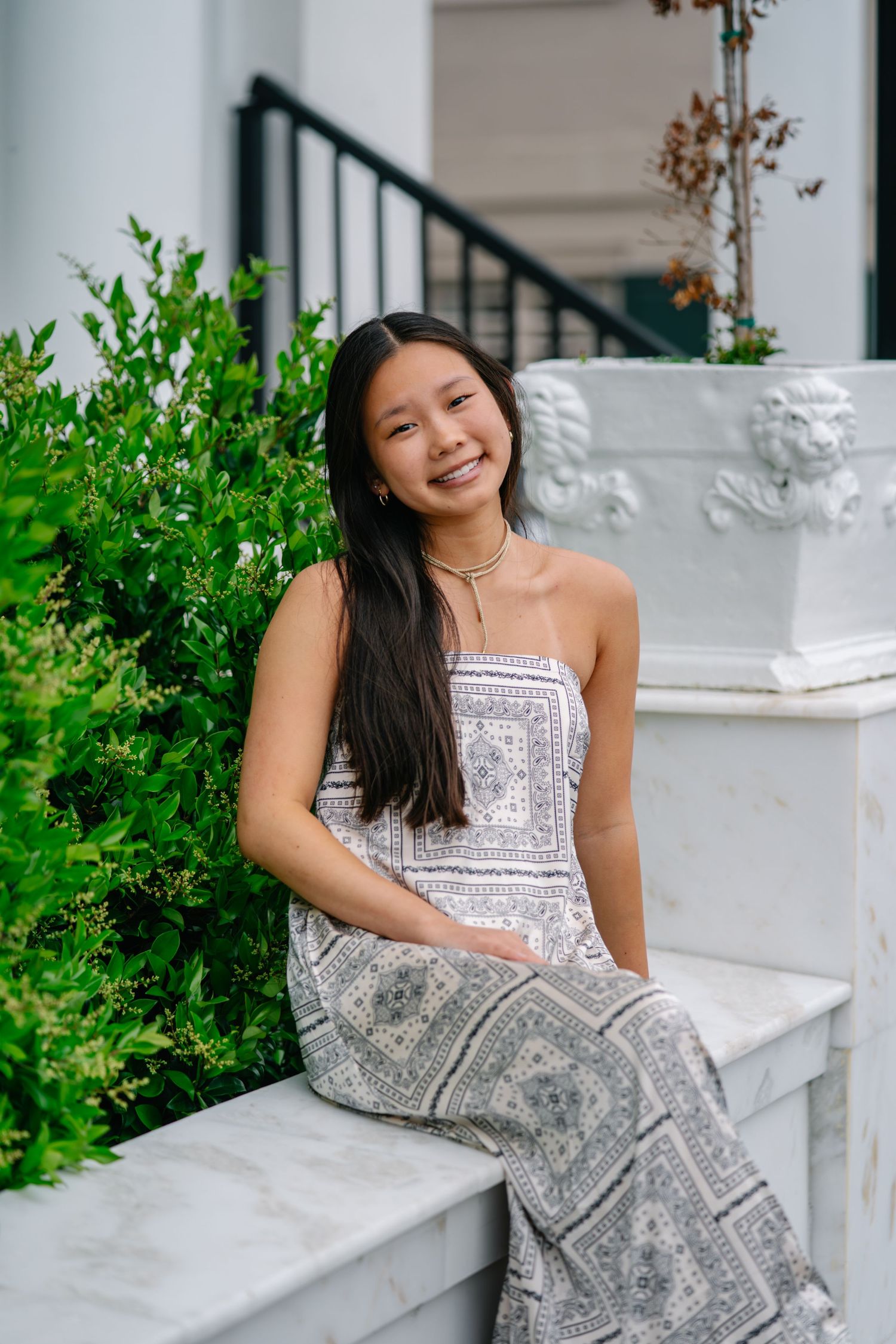 Young woman in patterned strapless dress sitting on white marble steps near green bushes, smiling outdoors.
