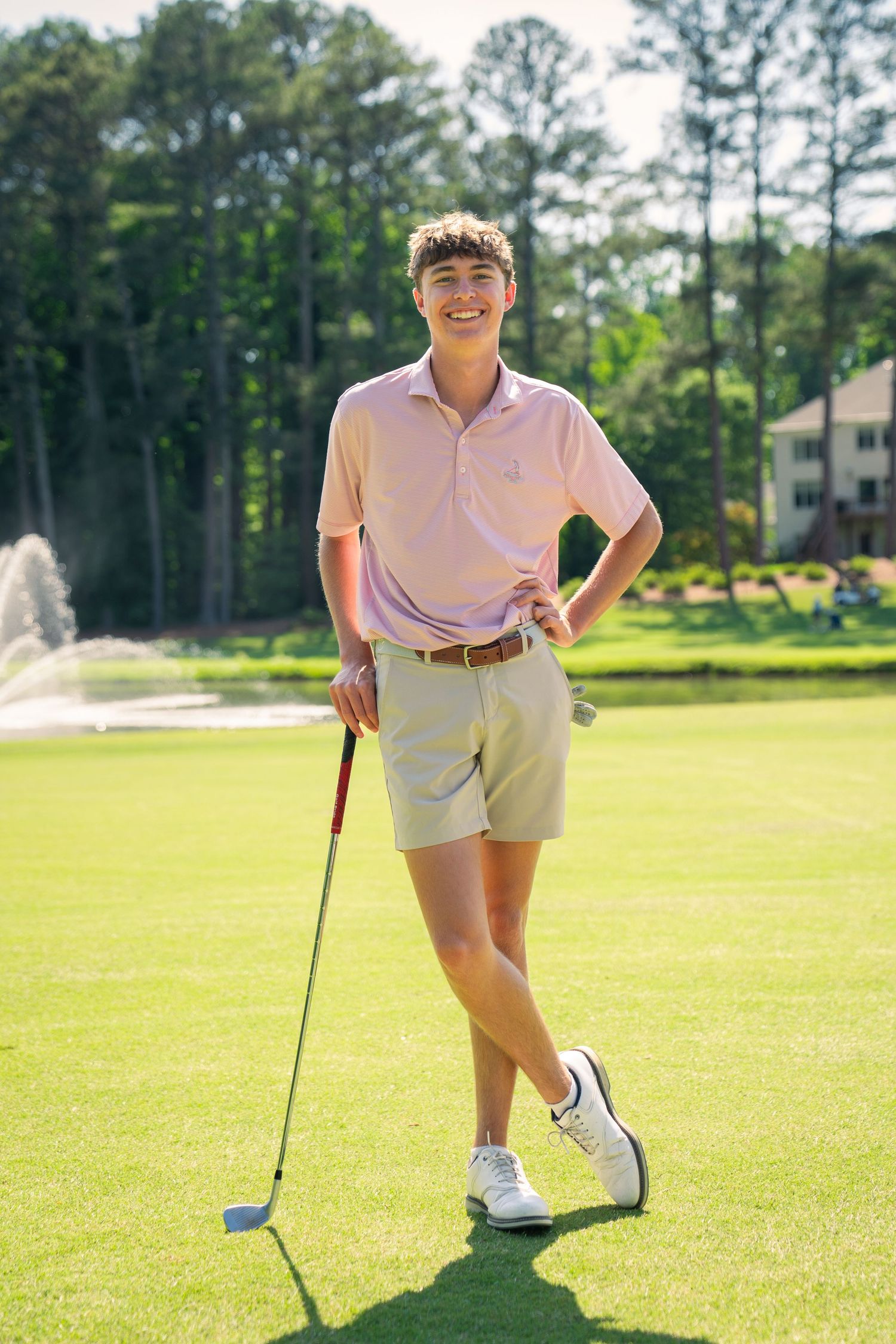 Young golfer in pink polo and khaki shorts poses with a golf club on a sunny green course with fountain backdrop.