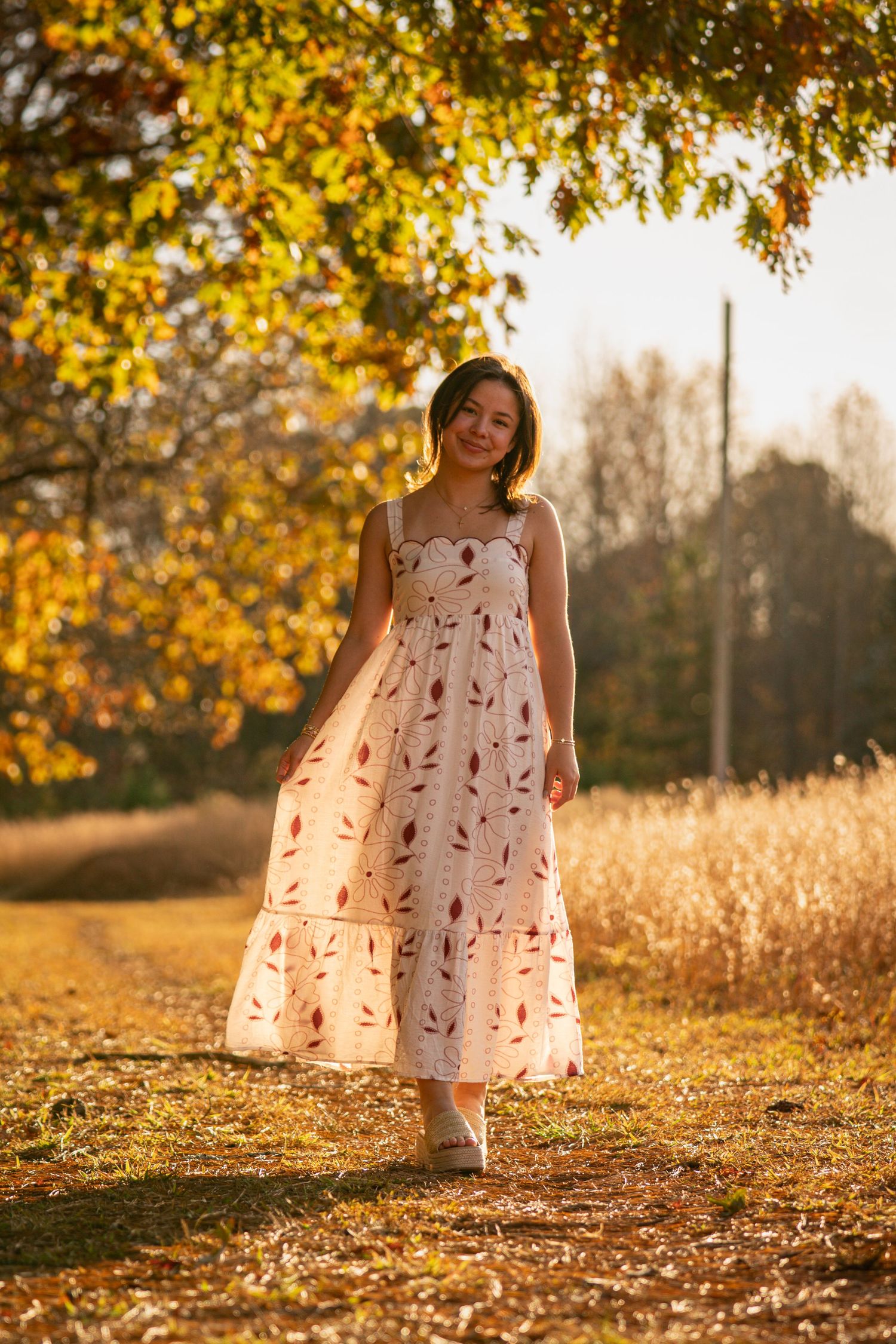 A person in a white floral dress stands in golden autumn sunlight under yellow leaves in a park setting.