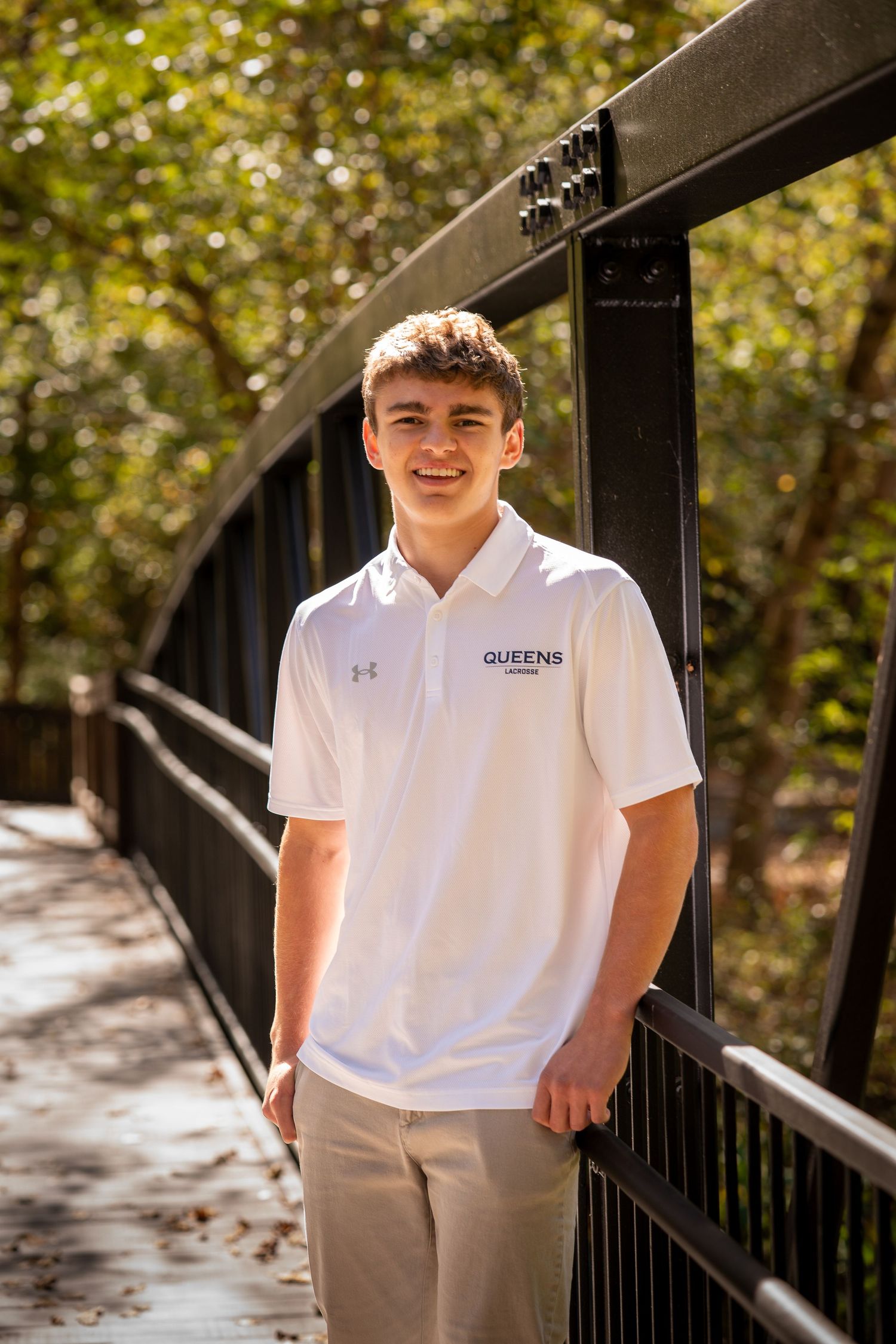 Student wearing white Queens polo shirt poses outdoors against black railing in autumn setting.