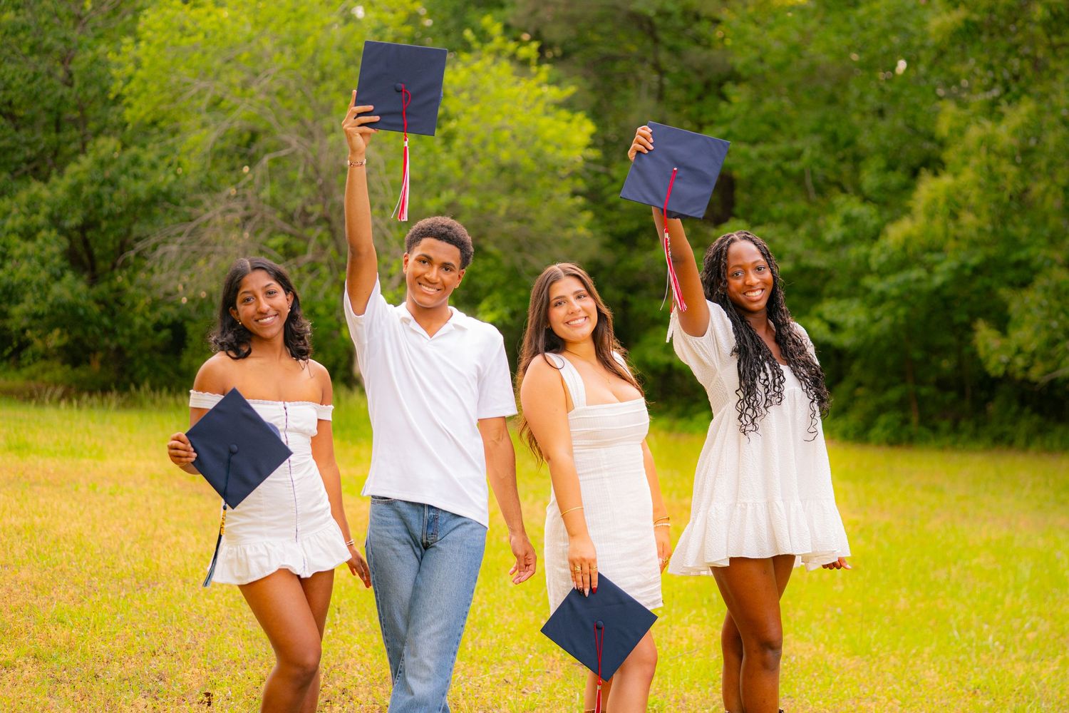 Four graduates in white dresses and a white shirt celebrate outdoors, holding navy graduation caps aloft in a green field.